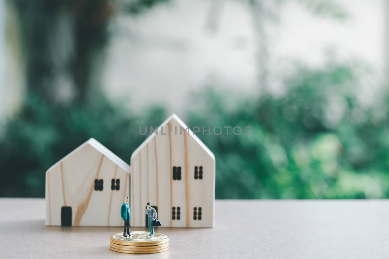 Saving money concept. Miniature people or small model businessman figure standing on coins stack with wooden home on green nature background.