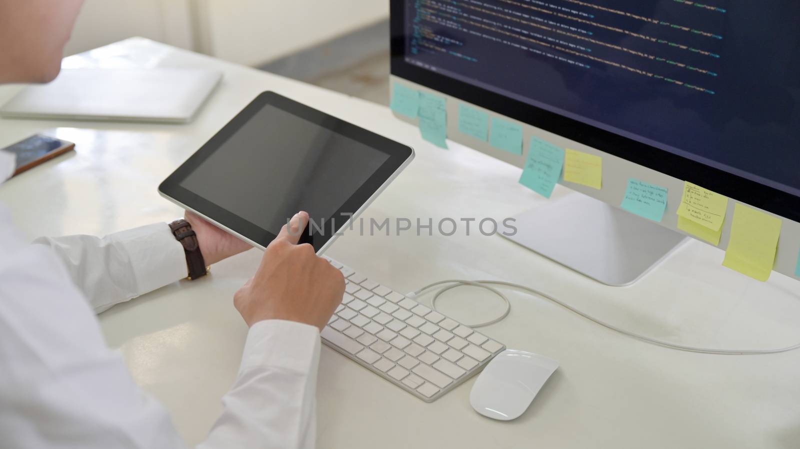 A programmer men using a tablet in his hand with a computer screen on his desk.