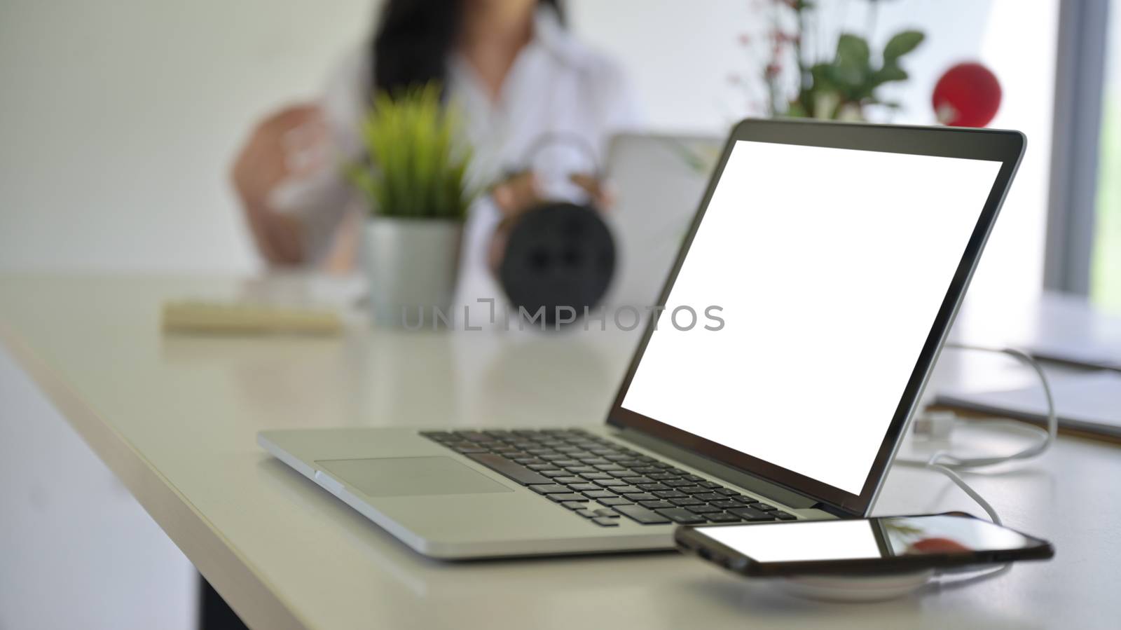 Laptop blank screen and smartphone on wireless charger with background people are working blurred.