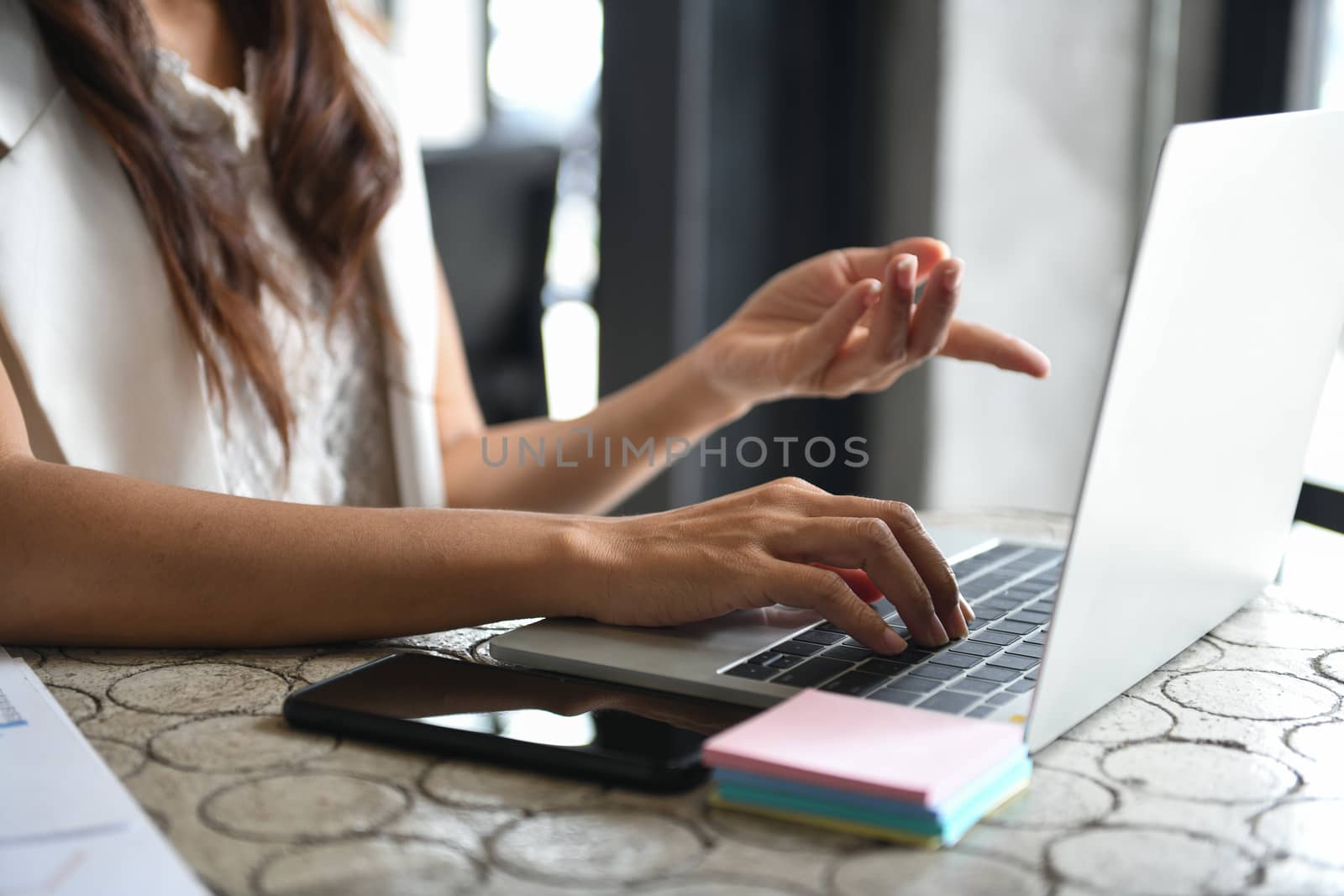 Cropped shot of a businesswoman using a laptop and having a smartphone on her side. She placed her hand on the keyboard and pointed her other hand at the screen.