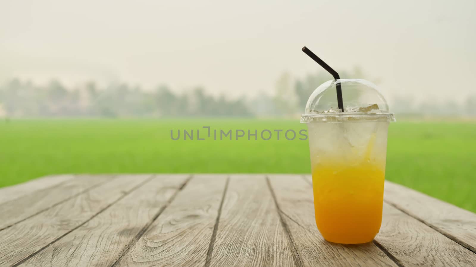Orange juice with ice in plastic mug on a wooden table with a natural green background, Cold drink for hot weather.