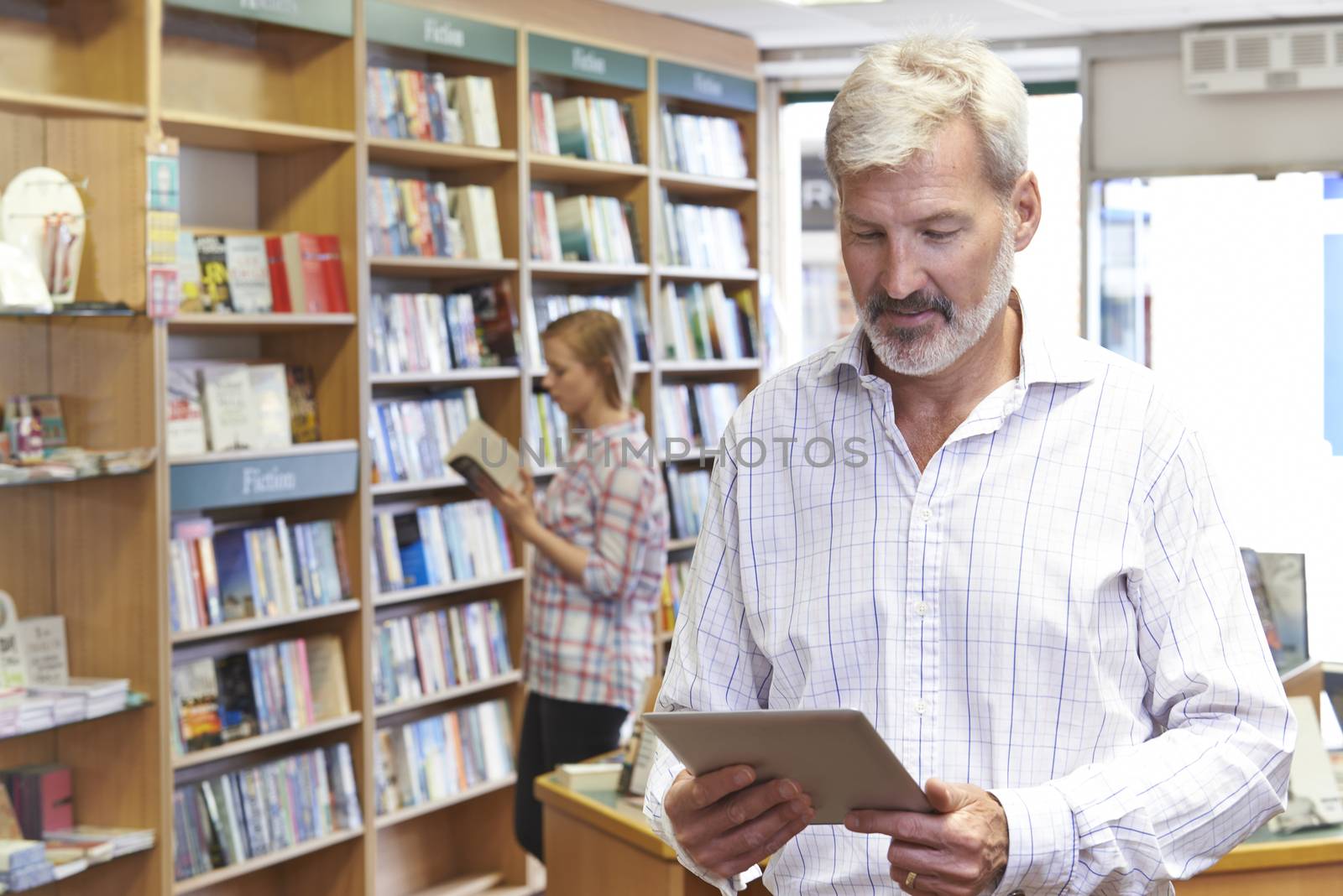 Male Bookstore Owner Using Digital Tablet With Customer In Backg by HWS