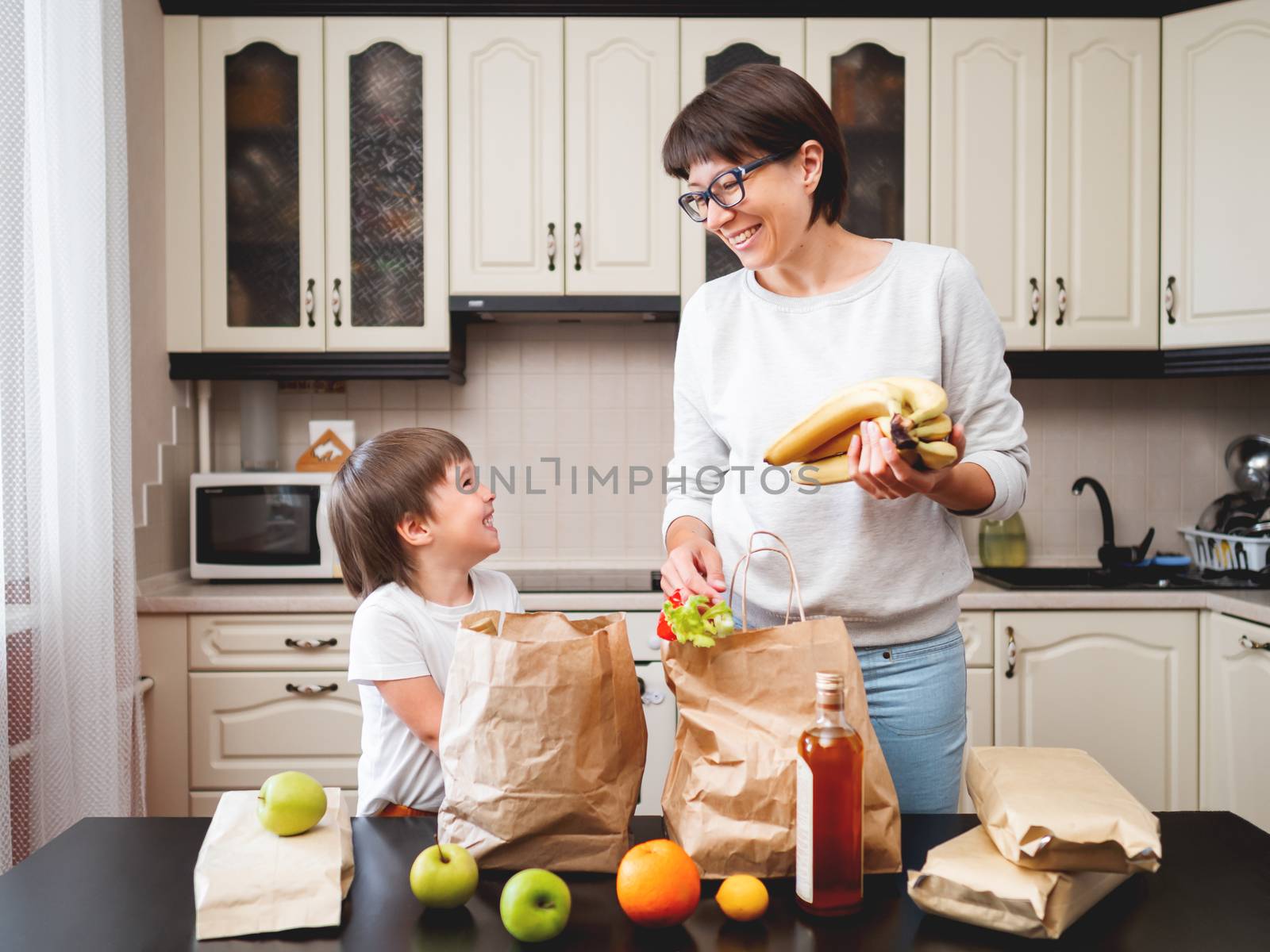 Woman and toddler boy sorts out purchases in the kitchen. Kid bites an apple. Grocery delivery in paper bags. Subscription service from grocery store. Mother and son at kitchen.