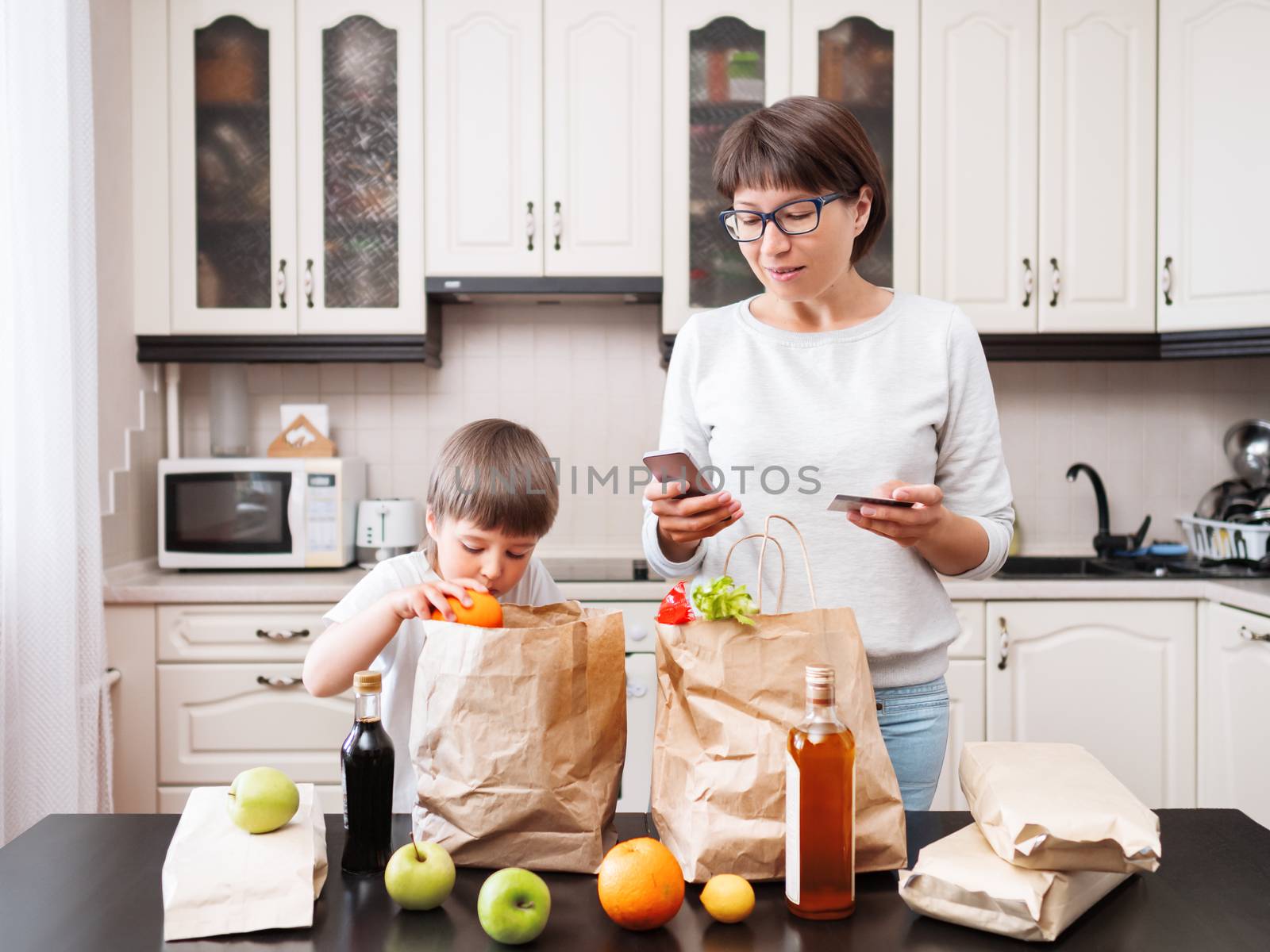 Woman and toddler boy sorts out purchases in the kitchen. Grocery delivery in paper bags. Subscription service from grocery store. Mother and son at kitchen.