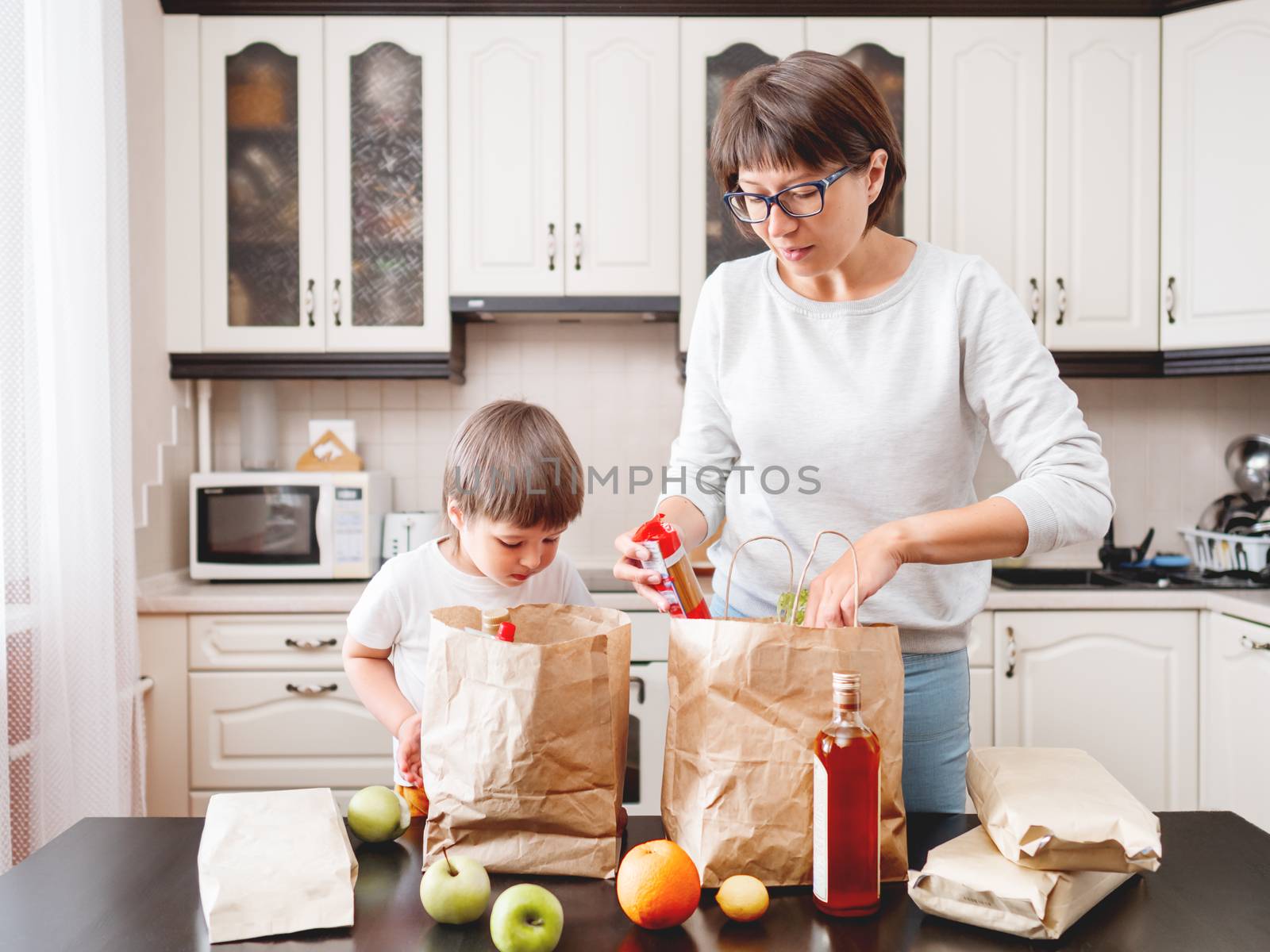 Woman and toddler boy sorts out purchases in the kitchen. Kid bites an apple. Grocery delivery in paper bags. Subscription service from grocery store. Mother and son at kitchen.