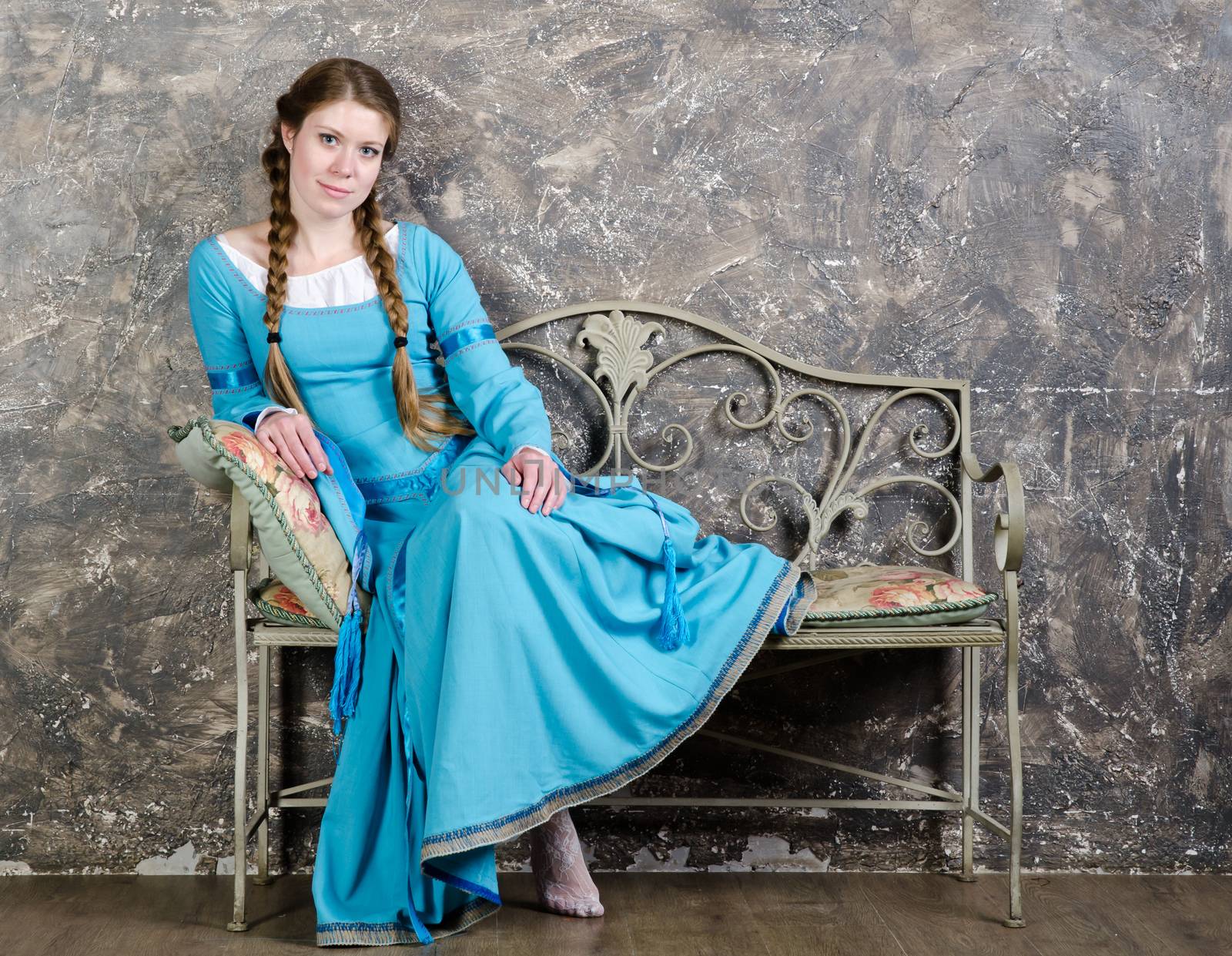 Playful young woman in historical medieval blue dress sits on a banquette