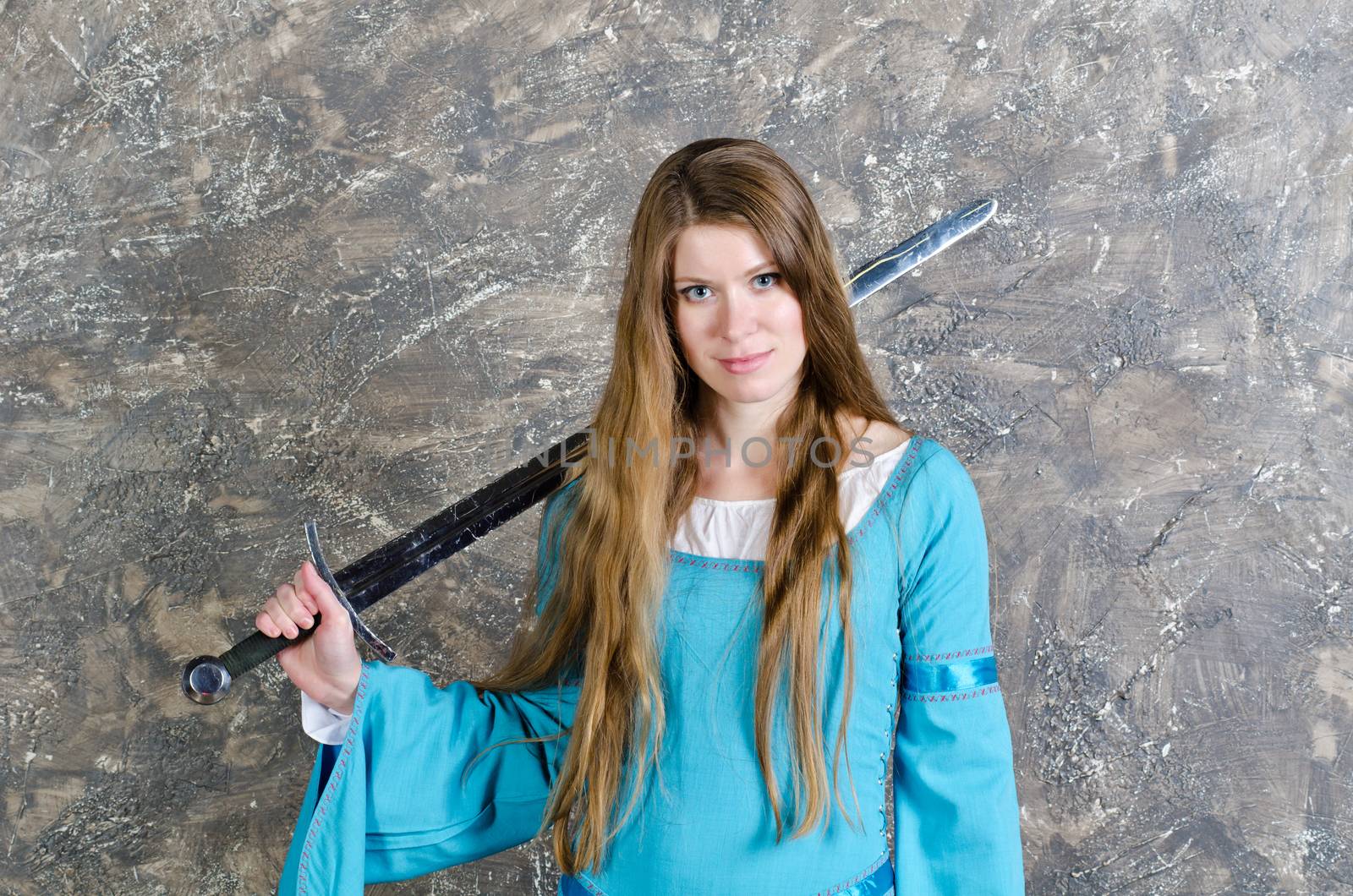 Pretty young woman with long hair in historical medieval blue dress poses in studio with sword