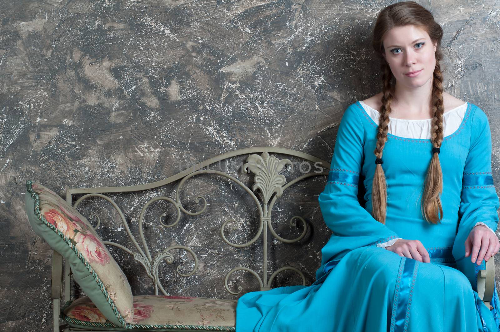 Pretty young woman in historical medieval blue dress sits on a banquette in studio