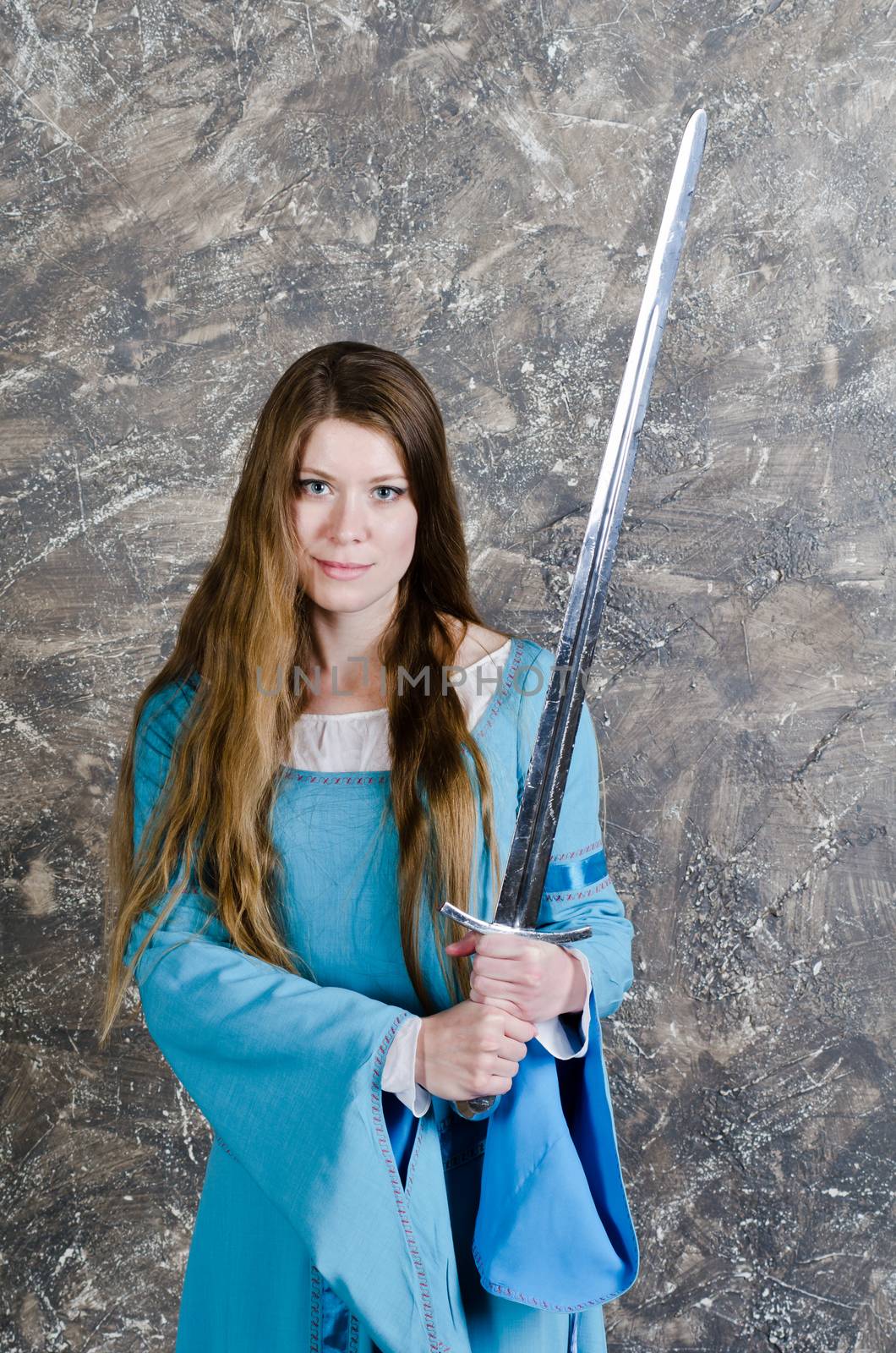 Pretty young woman with long hair in historical medieval blue dress poses in studio with sword