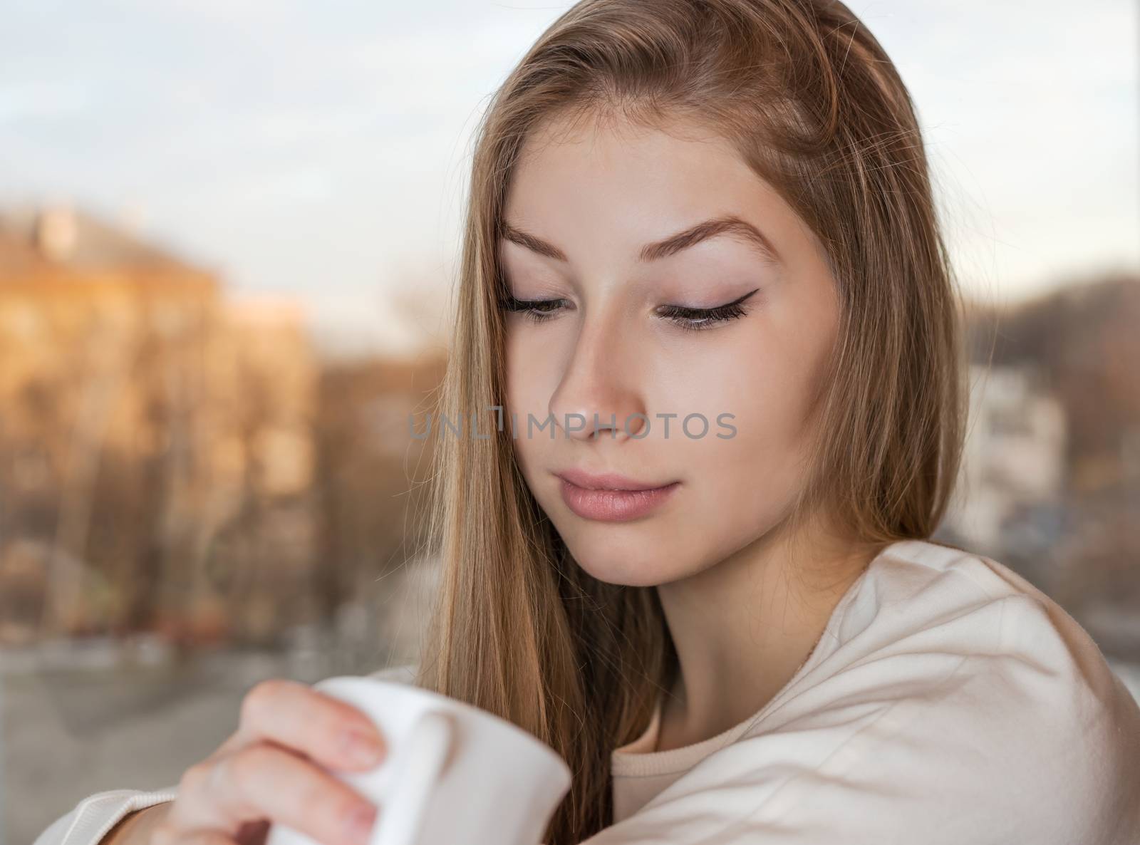 Lovely girl is sitting indoor near the window and enjoying the aroma from a white mug