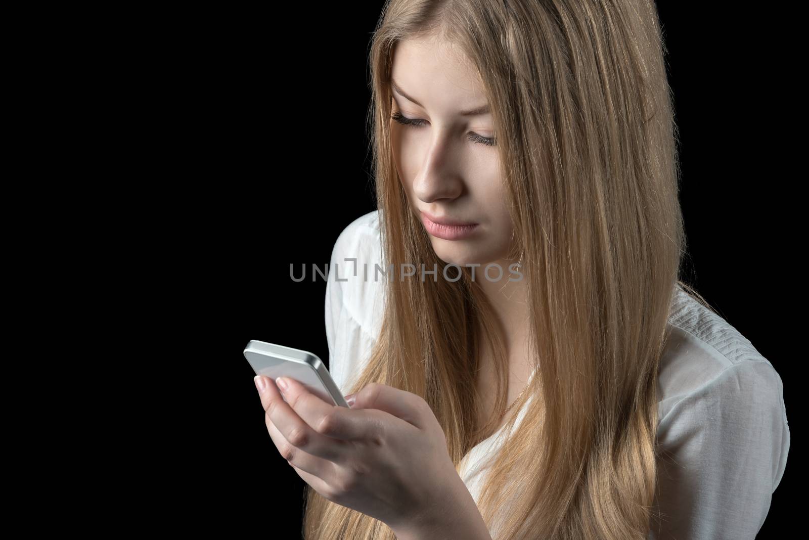 Attractive young girl feels depressed after reading bad sms on her mobile cellular phone. Isolated on black.