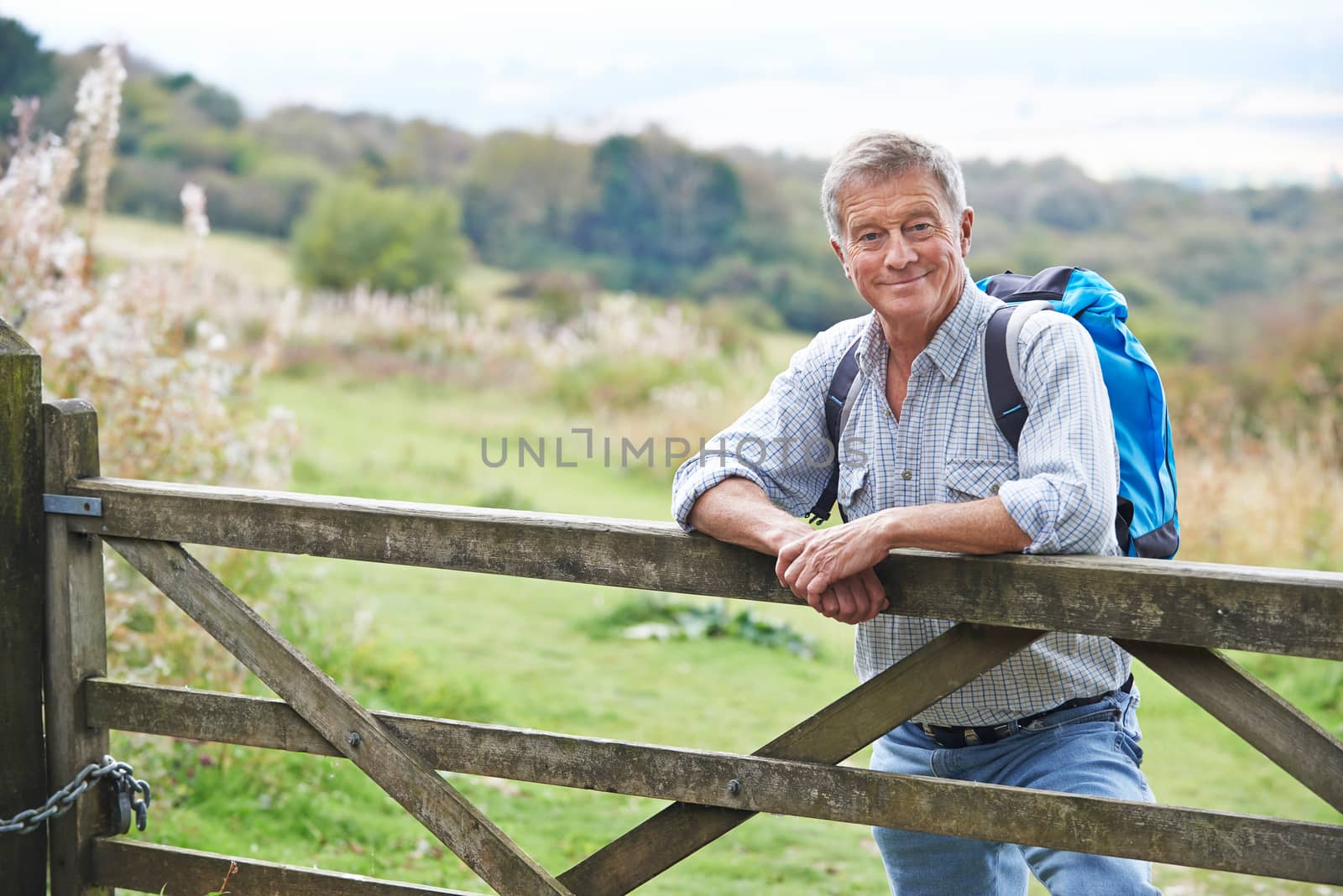 Portrait Of Senior Man On Hike In Countryside Resting By Gate by HWS