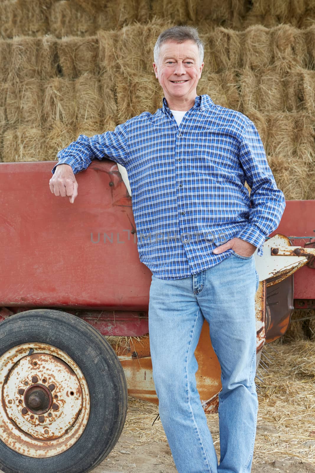 Farmer Standing In Front Of Straw Bales And Old Farm Equipment by HWS