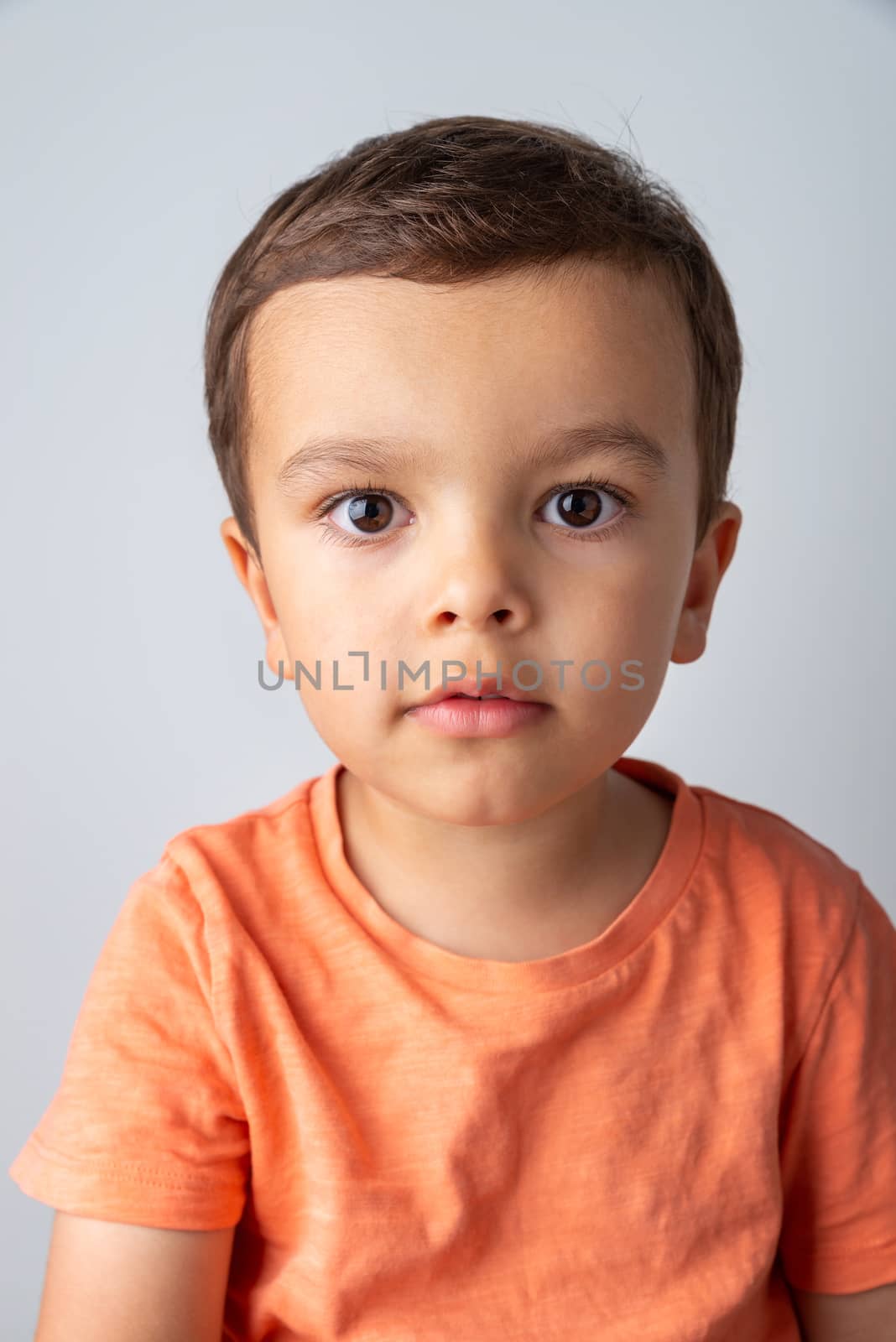 Cute three year old boy portrait, toddler wearing orange tee shirt and shot against a light grey background.