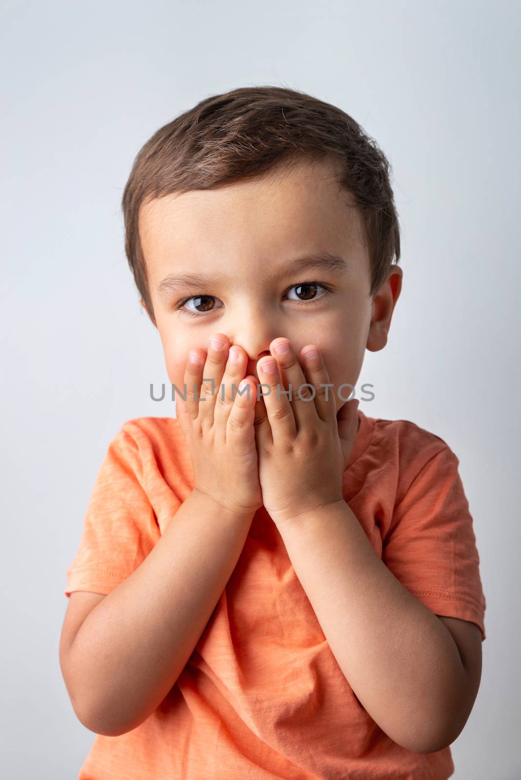 Cute three year old boy portrait, toddler covering his mouth with both hands.