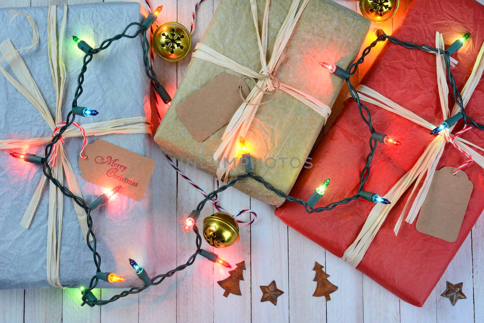 Three tissue wrapped Christmas presents on a rustic white wood table. A string of holiday lights and ornaments are draped over the packages. 