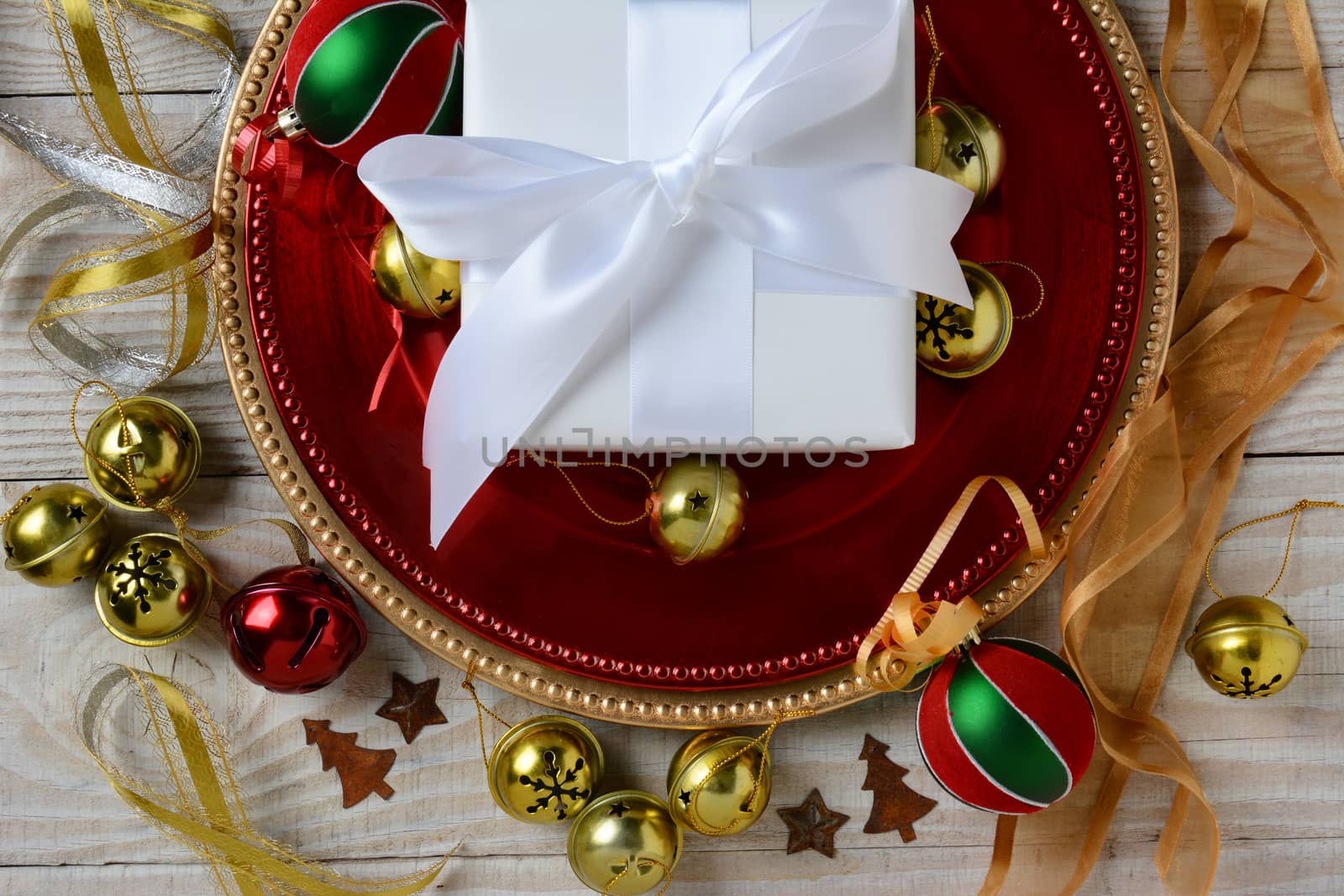 Overhead view of a white paper wrapped Christmas Present on red and gold chargers surrounded by holiday ornaments and ribbon. Horizontal format.