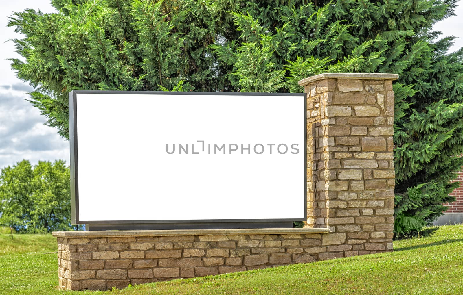 Horizontal shot of a big blank sign on a stonework support with grass and greenery around it.