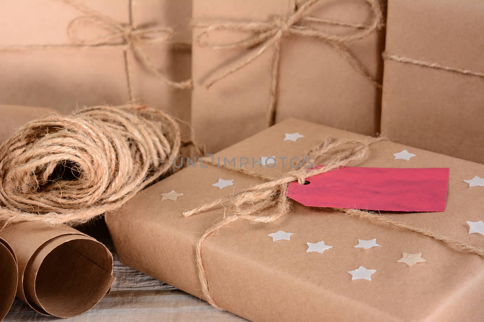 Closeup of a group of Christmas packages wrapped with plain brown paper and twine. Horizontal format on a wood table. A red gift tag and paper stars adorn one parcel.