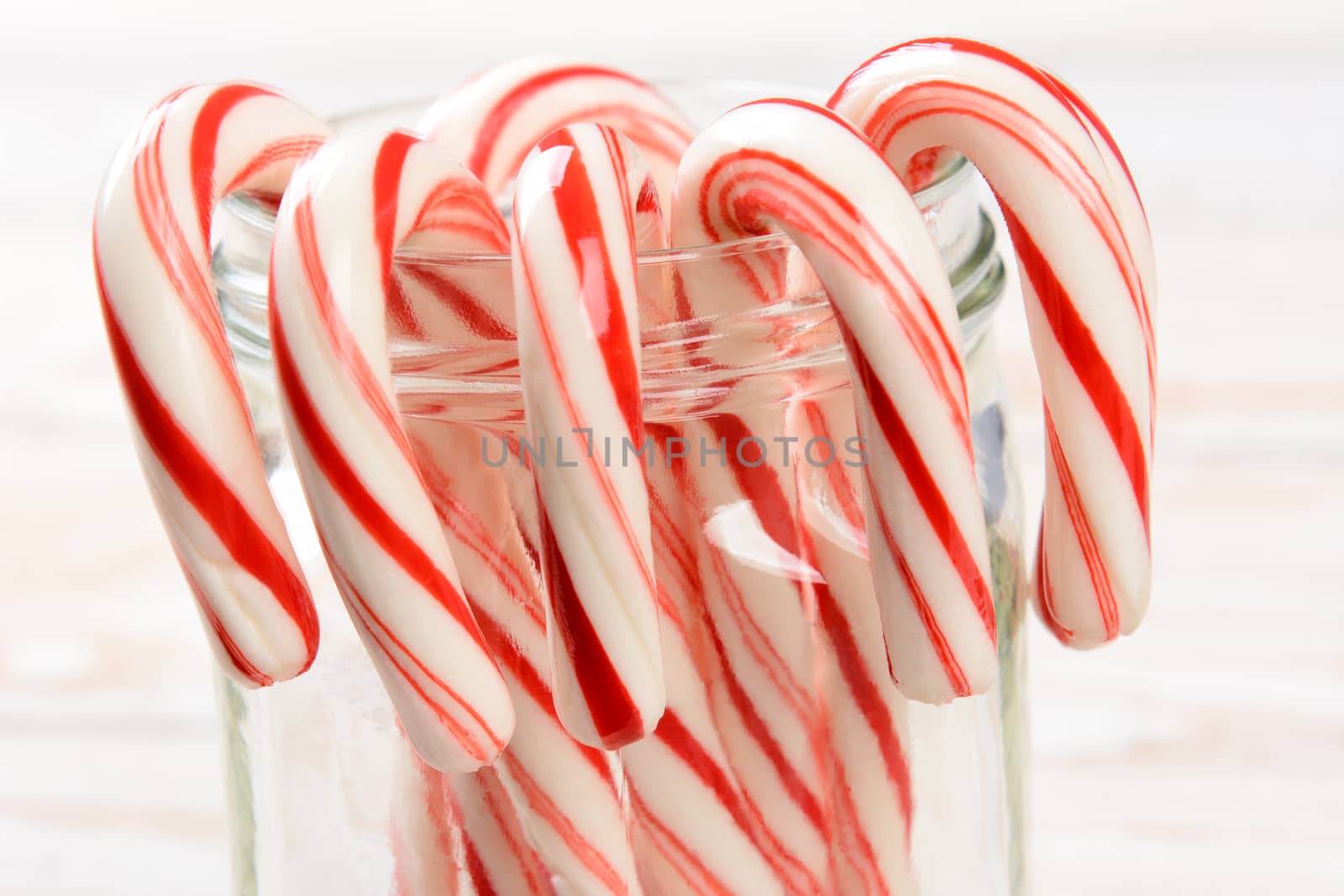 Closeup of a bunch of holiday candy canes hooked on the rim of a glass jar. Shallow depth of field on a whitewashed wood background. Horizontal format.