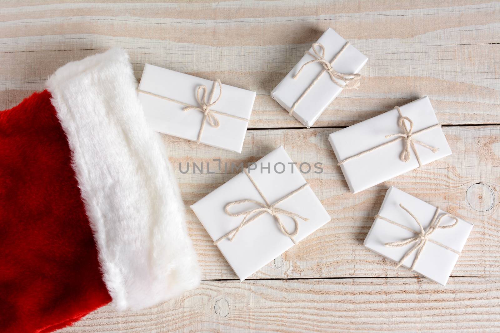 High angle photo of five Christmas presents wrapped in white paper and tied with white string and a stocking. The gifts are on a whitewashed wood table. Horizontal format.