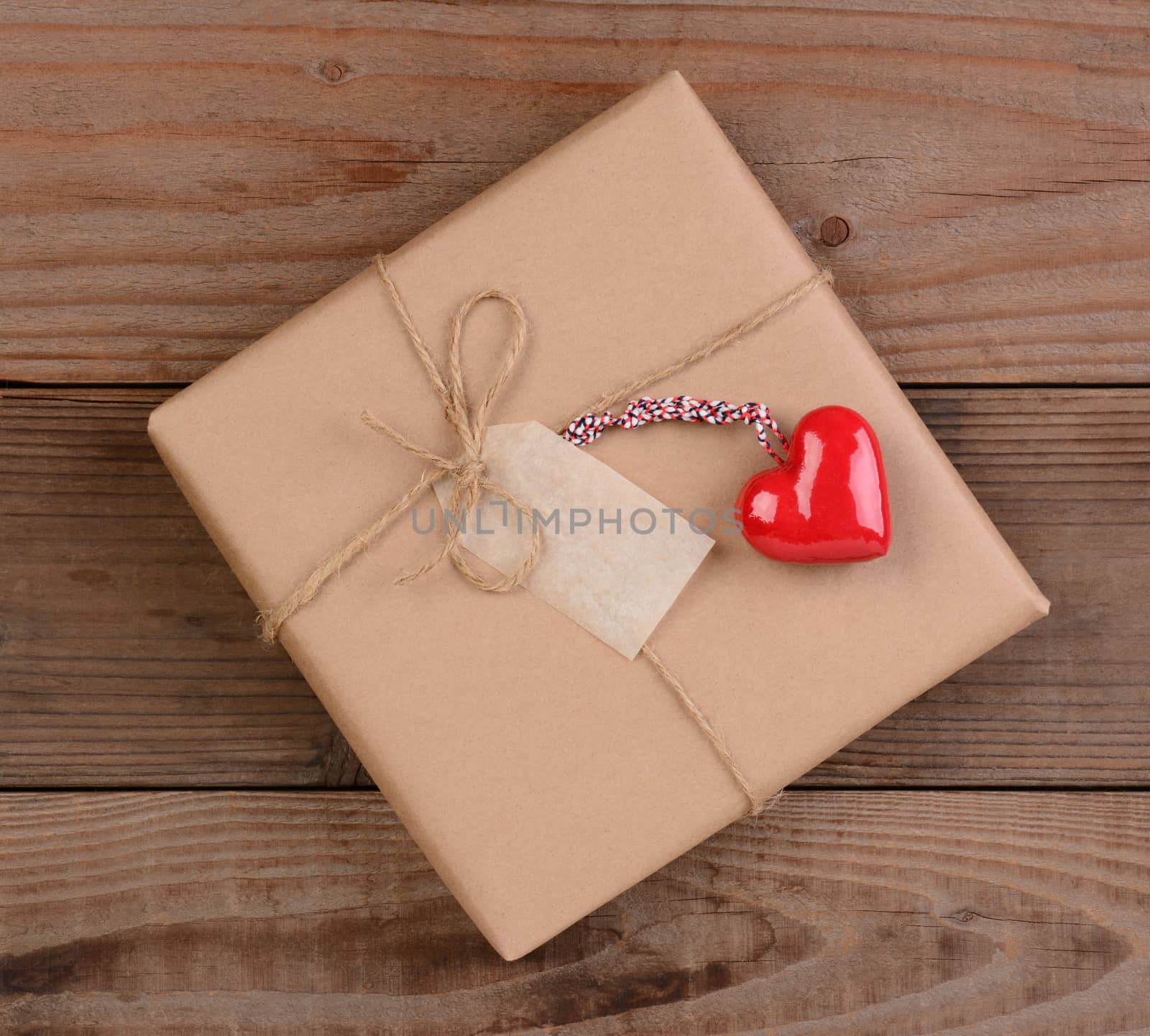 High angle view of a plain brown paper wrapped Valentines Day present with a ceramic red heart. Square format on a rustic wood surface.