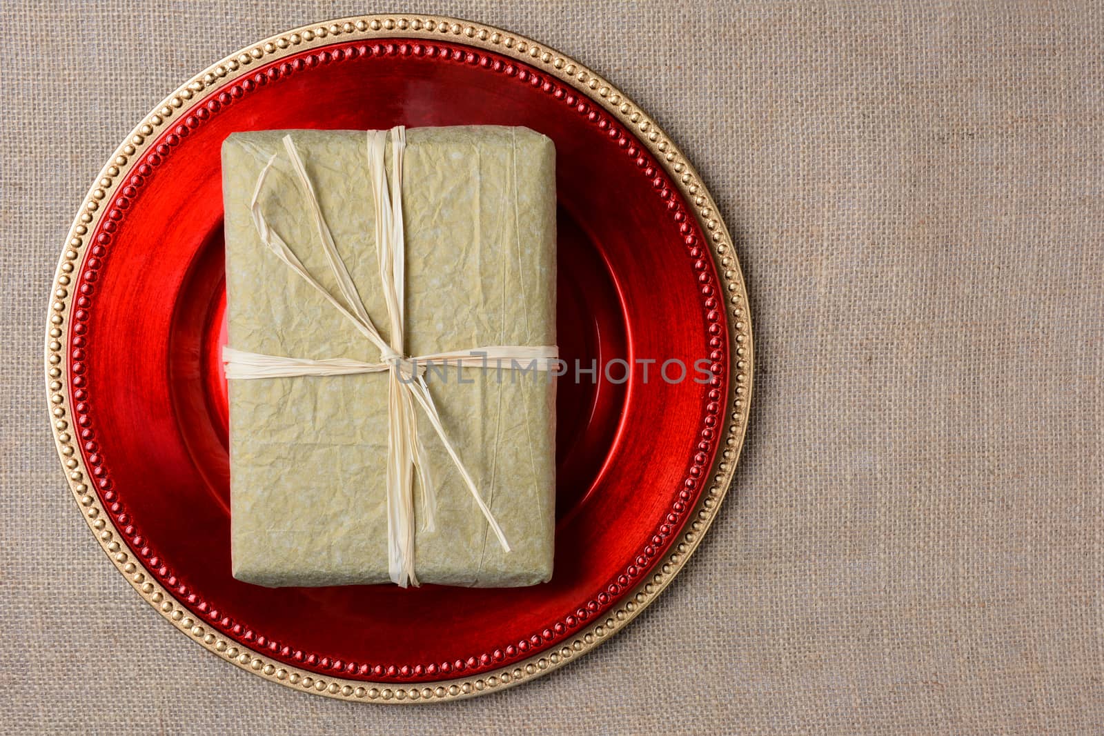 A gold tissue paper wrapped Christmas present on a red charger. The present is tied with raffia and both items are on a burlap surface. High angle shot in horizontal format with copyspace.