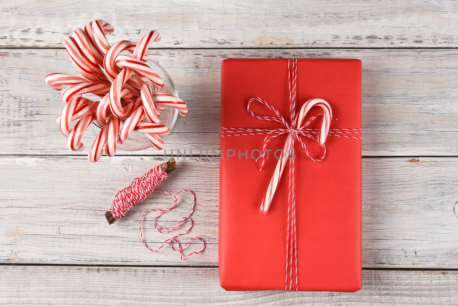 High angle view of a red paper wrapped Christmas present tied with string and a candy cane, next to a spool of twine and glass of candy canes.