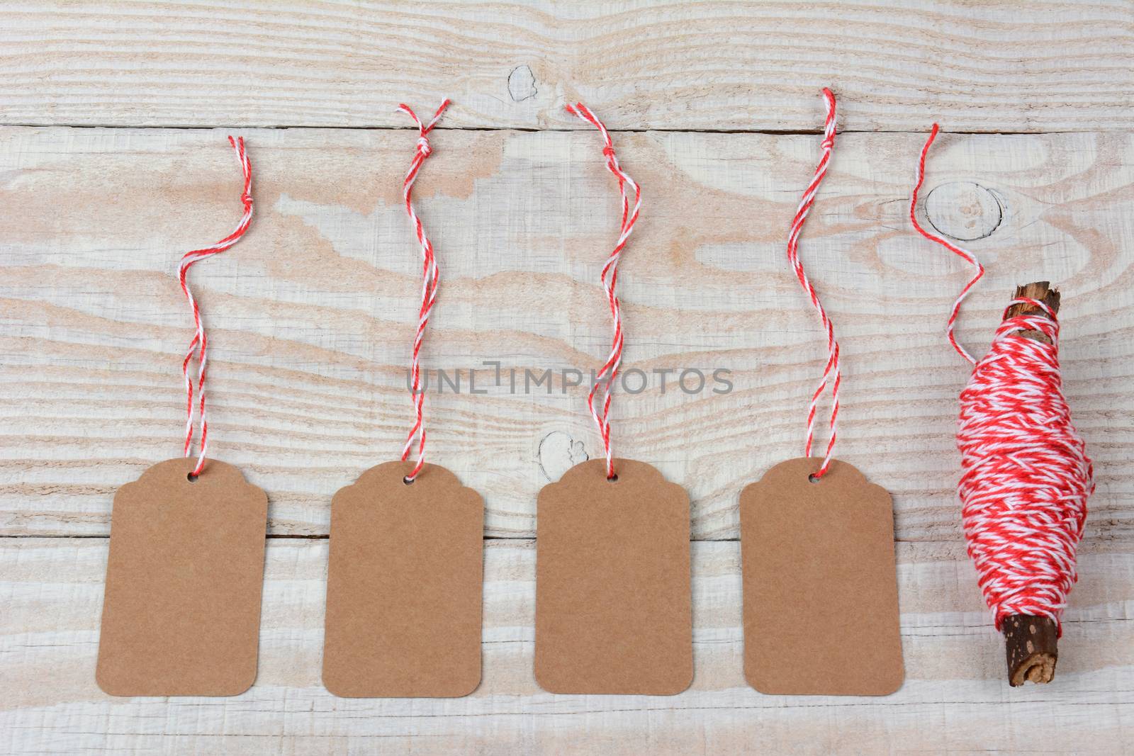 Overhead shot of four blank gift tags and red and white string wrapped around a twig laying on a rustic whitewashed wooden table. Horizontal format.