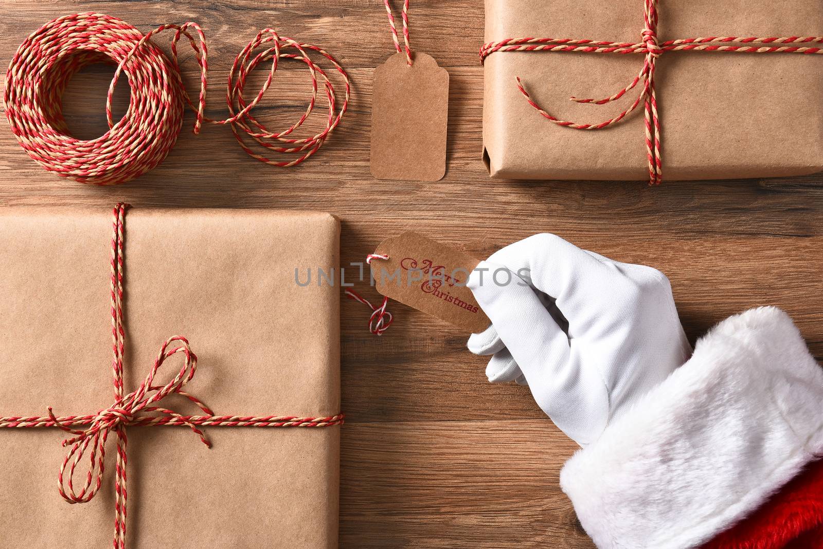 High angle view of Santa Claus hand holding a gift tag with Merry Christmas. Closeup with wrapped presents and string.