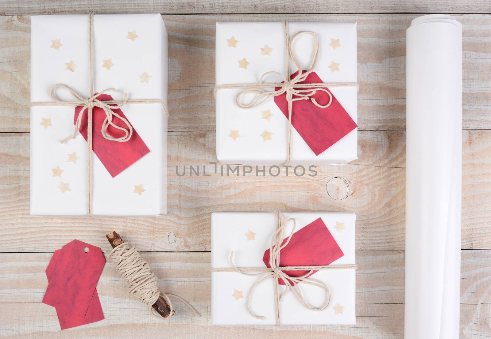 High angle shot of three Christmas presents wrapped in white paper and tied with white string. The gifts are on a white wood table with string, tags and a roll of paper.