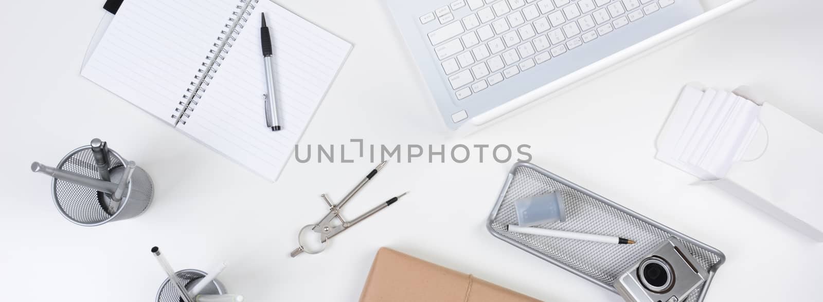 High angle shot of a white desk with primarily white and silver office objects. Items include, laptop, computer, pad, pens, camera, with a brown wrapped package at the bottom of the frame. Banner Format.