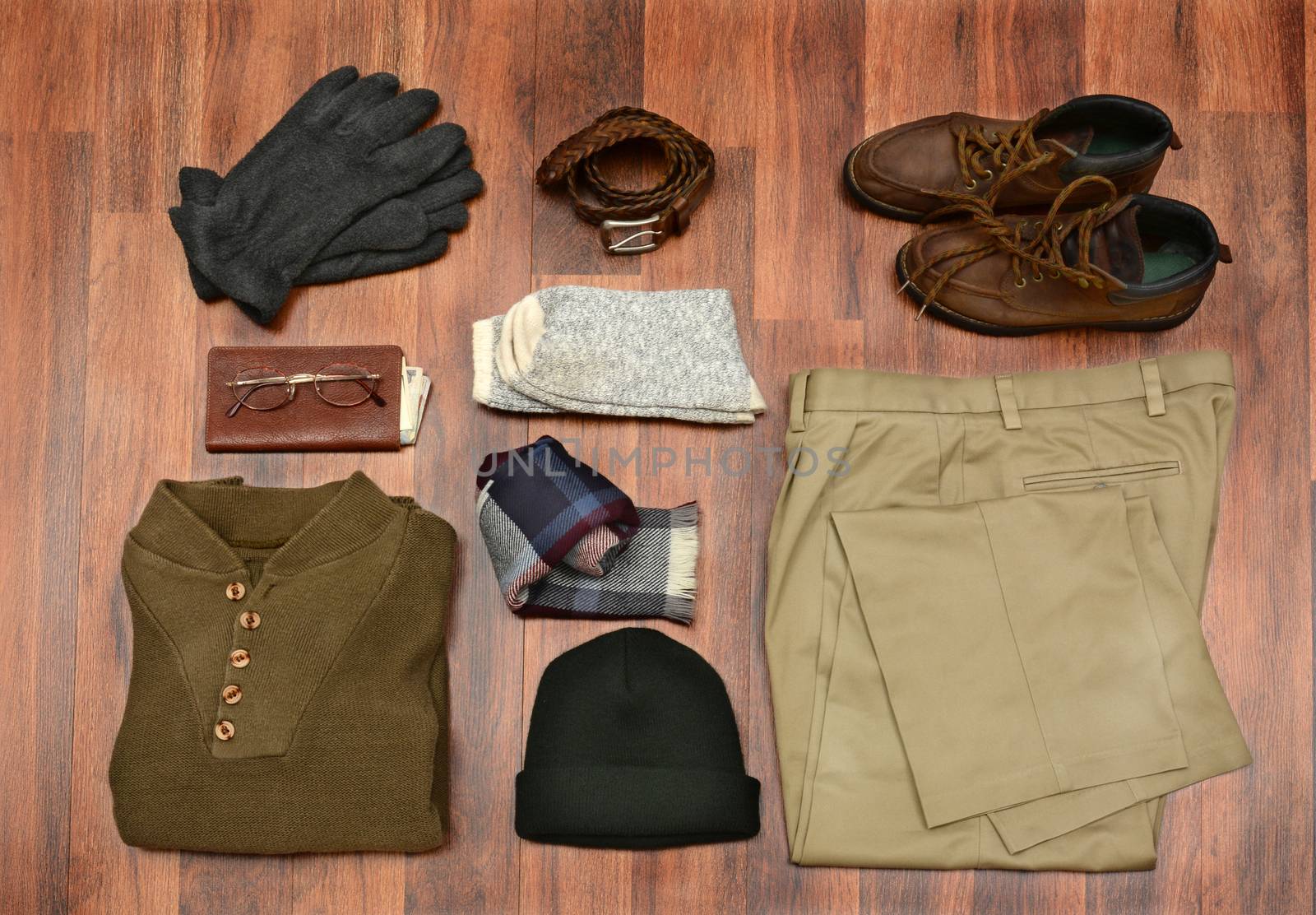High angle shot of men's winter clothes laid out on a dark wood floor. Items include, Sweater, Scarf, Gloves, wool Socks, Pants, Boots, belt, Knit Cap, Wallet, and Glasses. Horizontal Format.