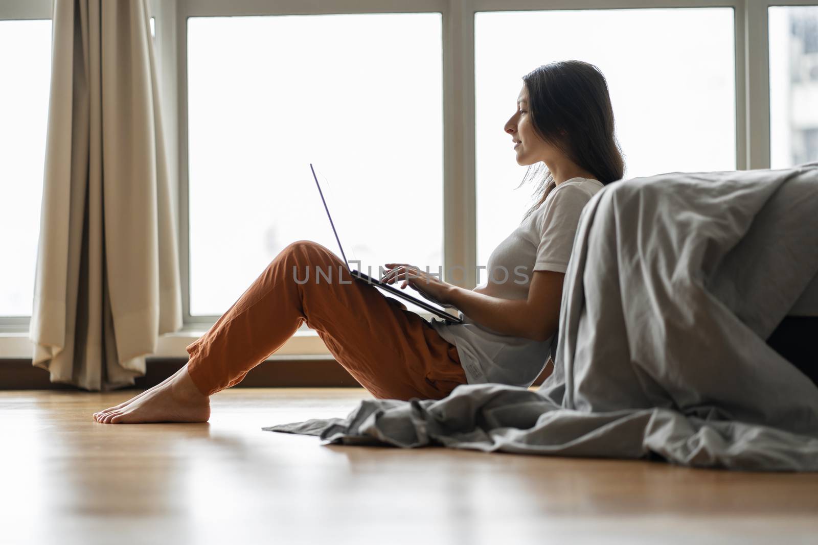 Beautiful young brunette girl working on a laptop, sitting on the floor near the bed by the panoramic window. Stylish modern interior. A cozy workplace. Shopping on the Internet by Try_my_best
