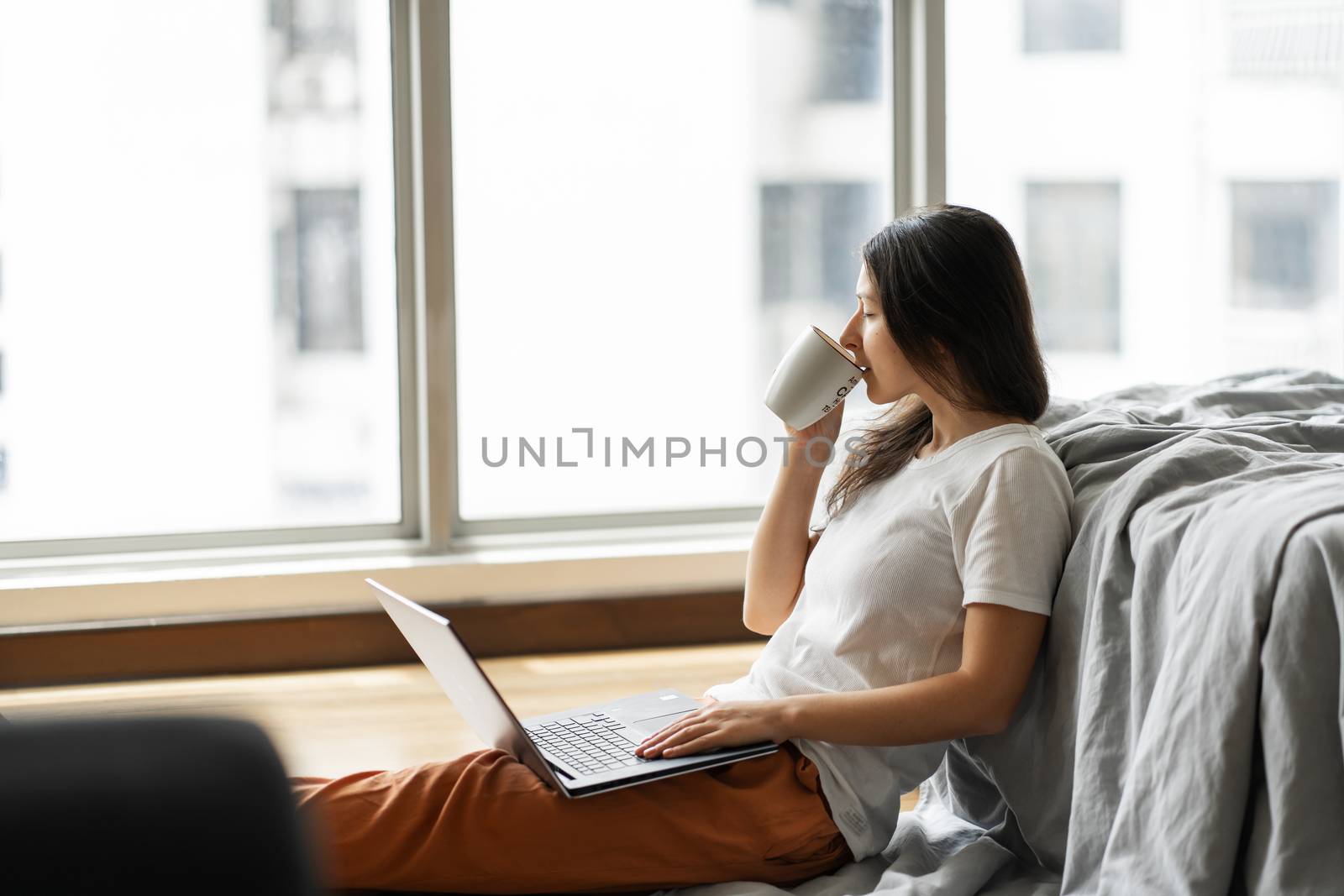 Beautiful young brunette girl working on a laptop and drinking coffee, sitting on the floor near the bed by the panoramic window. Stylish modern interior. A cozy workplace. Shopping on the Internet by Try_my_best