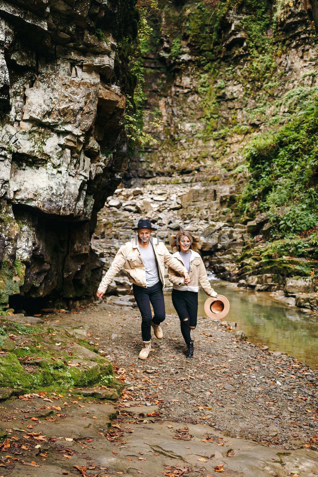 Young and beautiful couple at the mountain waterfall - Happy tourists visiting mountains. Lovestory. Tourists in hats. Military fashion. by Denys_N