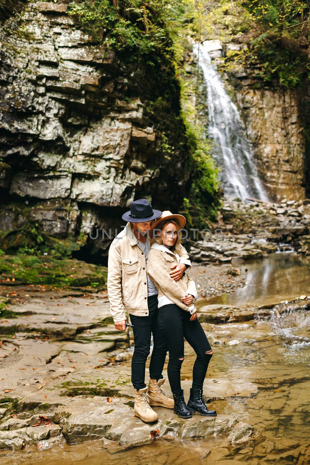 Young and beautiful couple at the mountain waterfall - Happy tourists visiting mountains. Lovestory. Tourists in hats. Military fashion. by Denys_N