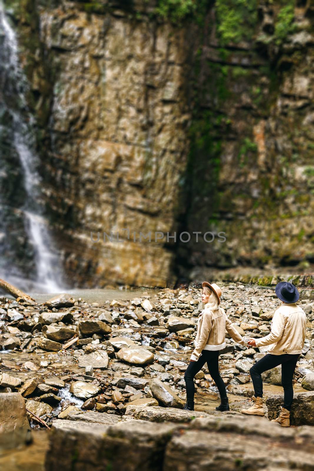 Young and beautiful couple at the mountain waterfall - Happy tourists visiting mountains. Lovestory. Tourists in hats. Military fashion. by Denys_N