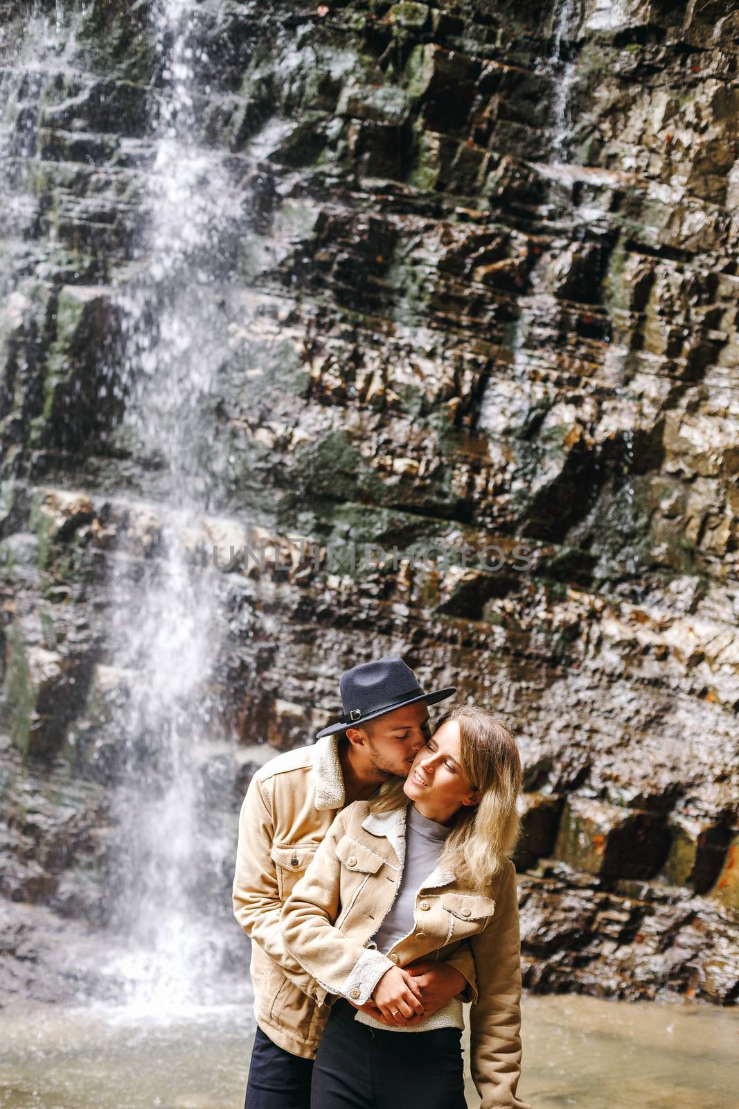 Young and beautiful couple at the mountain waterfall - Happy tourists visiting mountains. Lovestory. Tourists in hats. Military fashion. by Denys_N