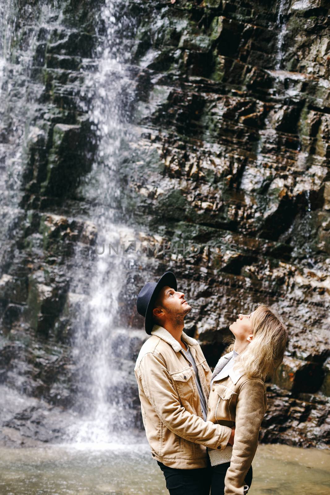 Young and beautiful couple at the mountain waterfall - Happy tourists visiting mountains. Lovestory. Tourists in hats. Military fashion. by Denys_N