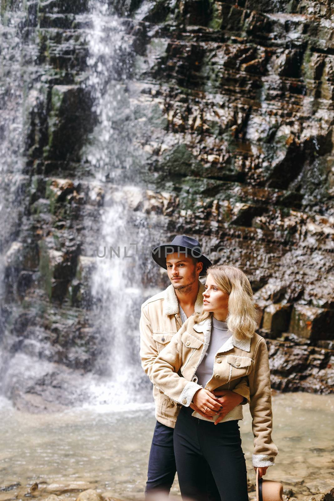 Young and beautiful couple at the mountain waterfall - Happy tourists visiting mountains. Lovestory. Tourists in hats. Military fashion. by Denys_N