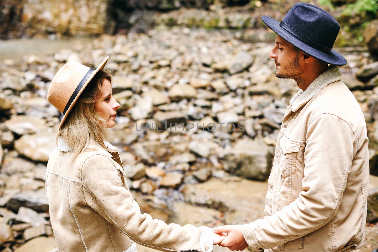 Young and beautiful couple at the mountain waterfall - Happy tourists visiting mountains. Lovestory. Tourists in hats. Military fashion. by Denys_N