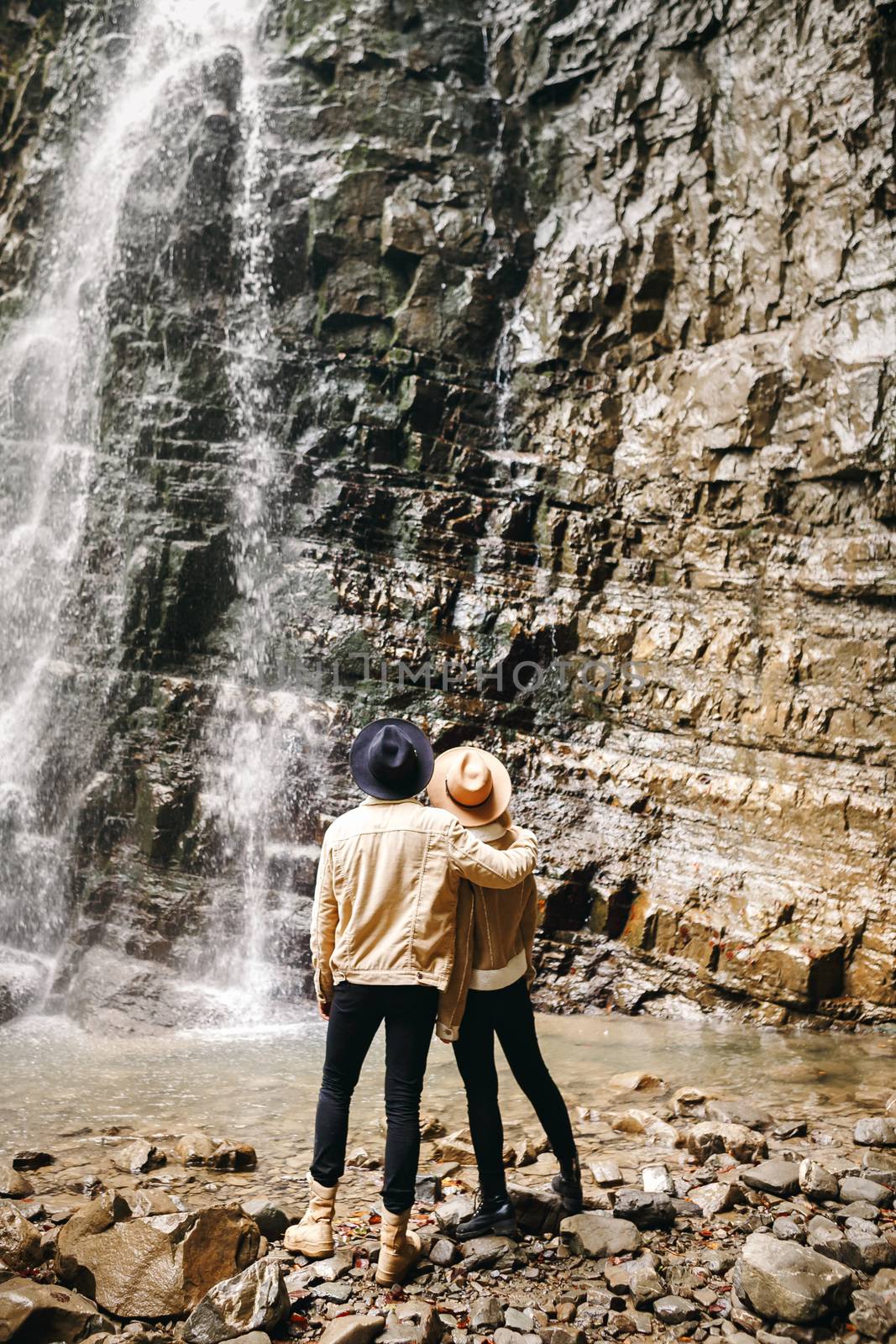 Young and beautiful couple at the mountain waterfall - Happy tourists visiting mountains. Lovestory. Tourists in hats. Military fashion