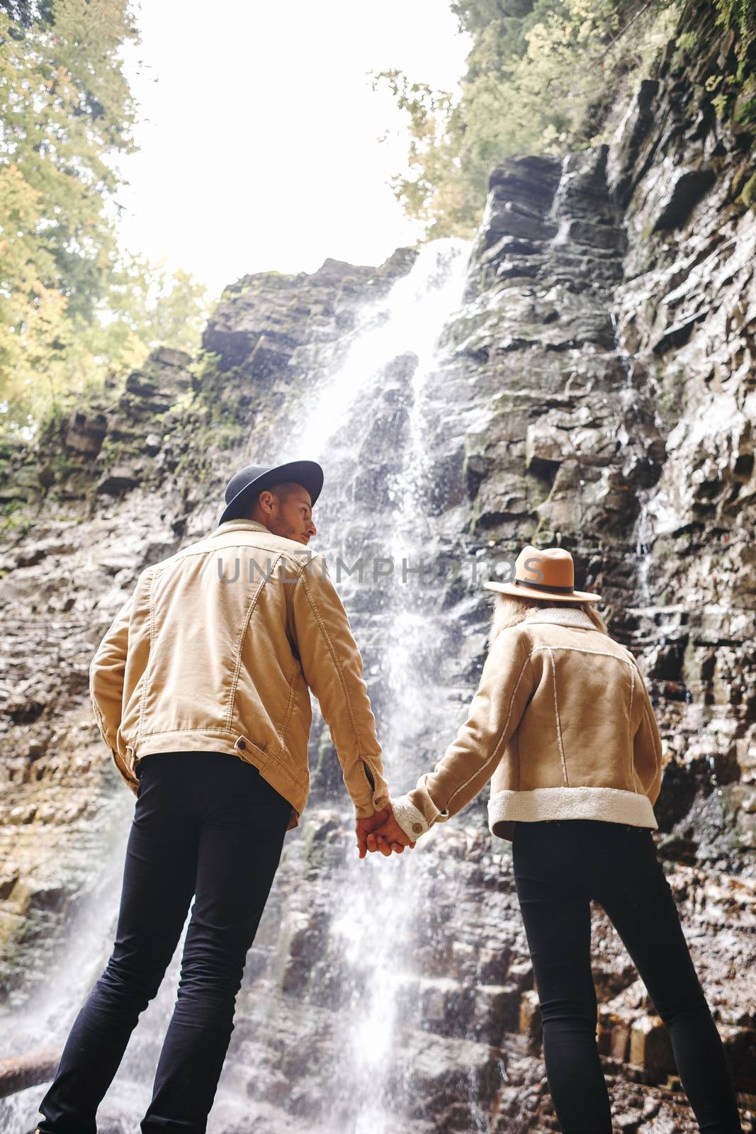 Young and beautiful couple at the mountain waterfall - Happy tourists visiting mountains. Lovestory. Tourists in hats. Military fashion. by Denys_N