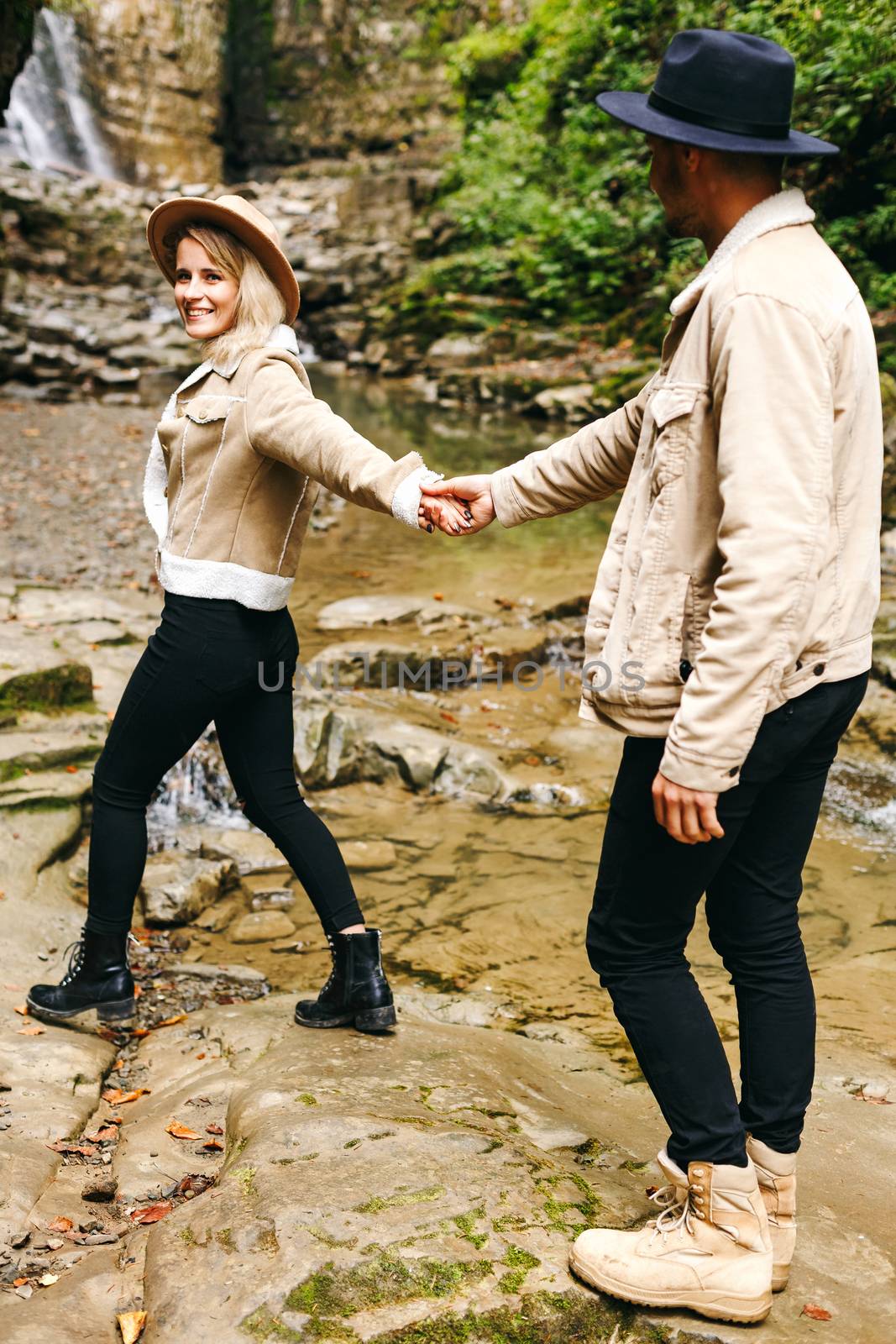 Young and beautiful couple at the mountain waterfall - Happy tourists visiting mountains. Lovestory. Tourists in hats. Military fashion. by Denys_N