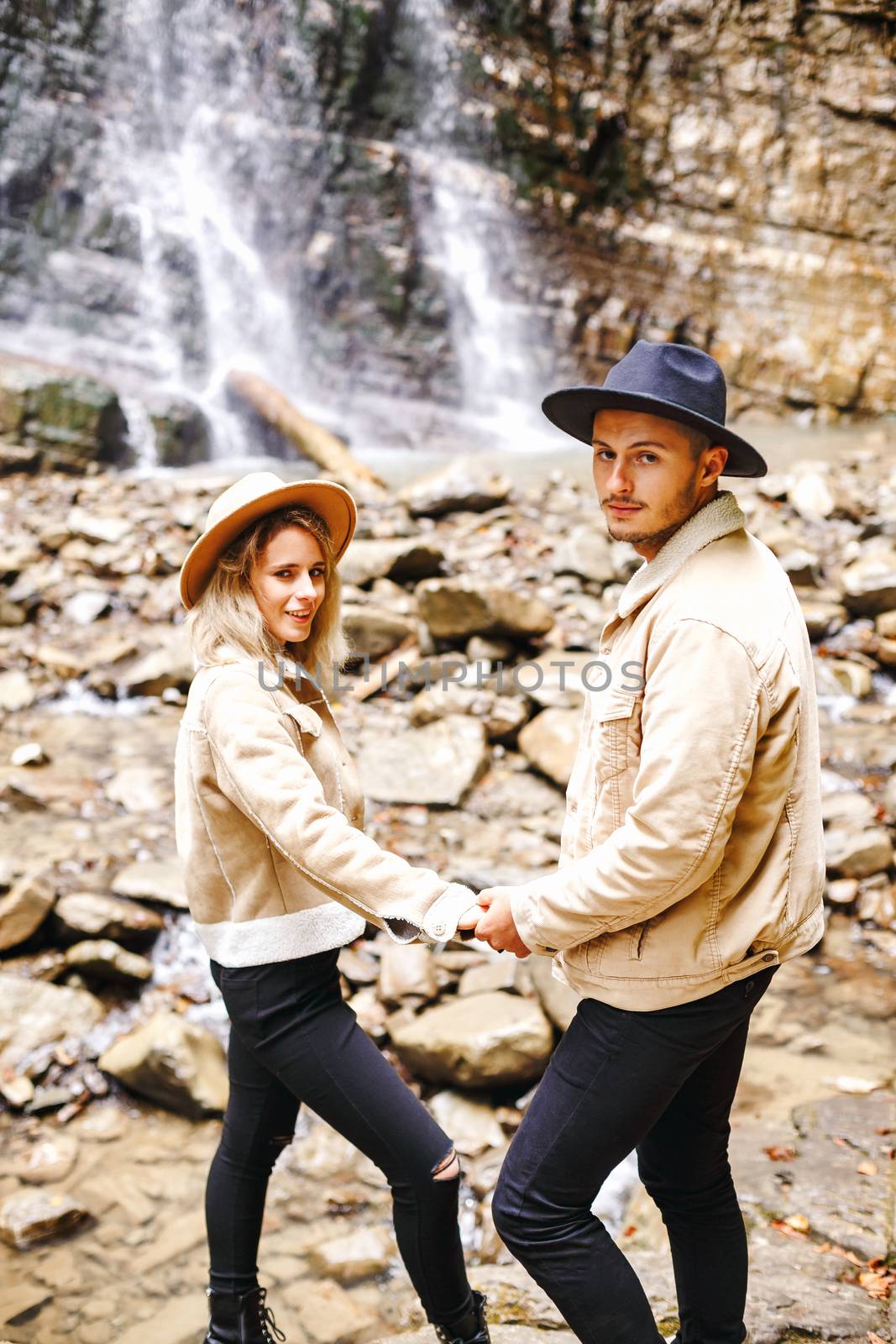 Young and beautiful couple at the mountain waterfall - Happy tourists visiting mountains. Lovestory. Tourists in hats. Military fashion. by Denys_N