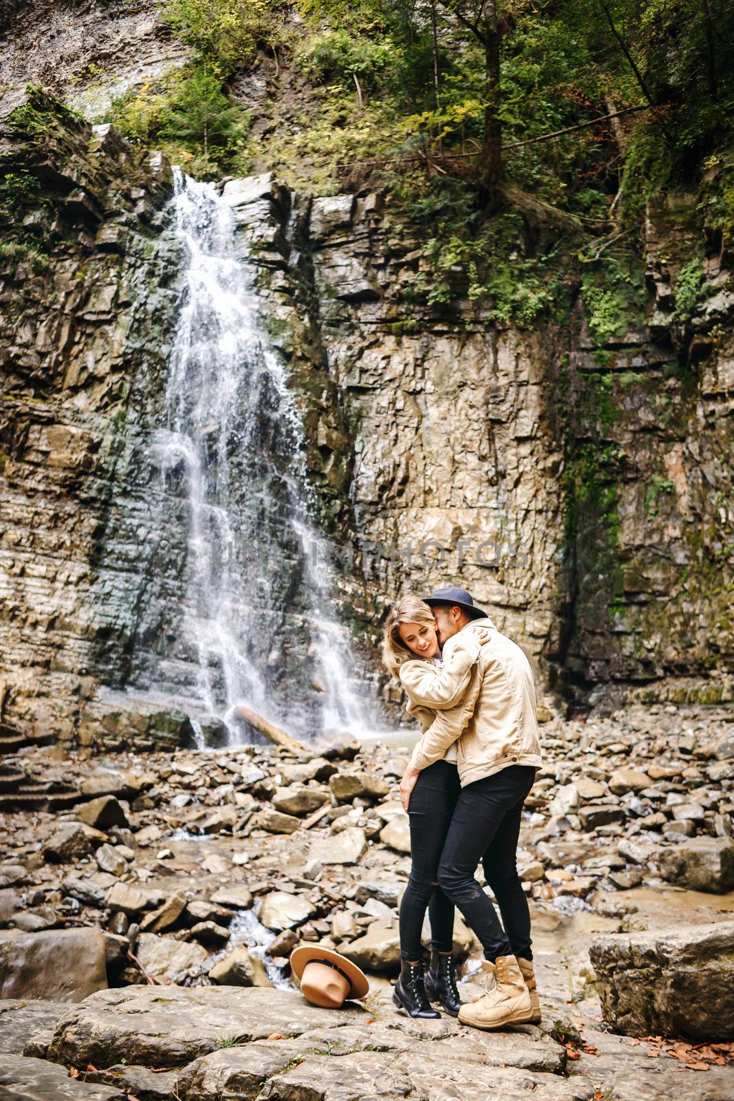 Young and beautiful couple at the mountain waterfall - Happy tourists visiting mountains. Lovestory. Tourists in hats. Military fashion