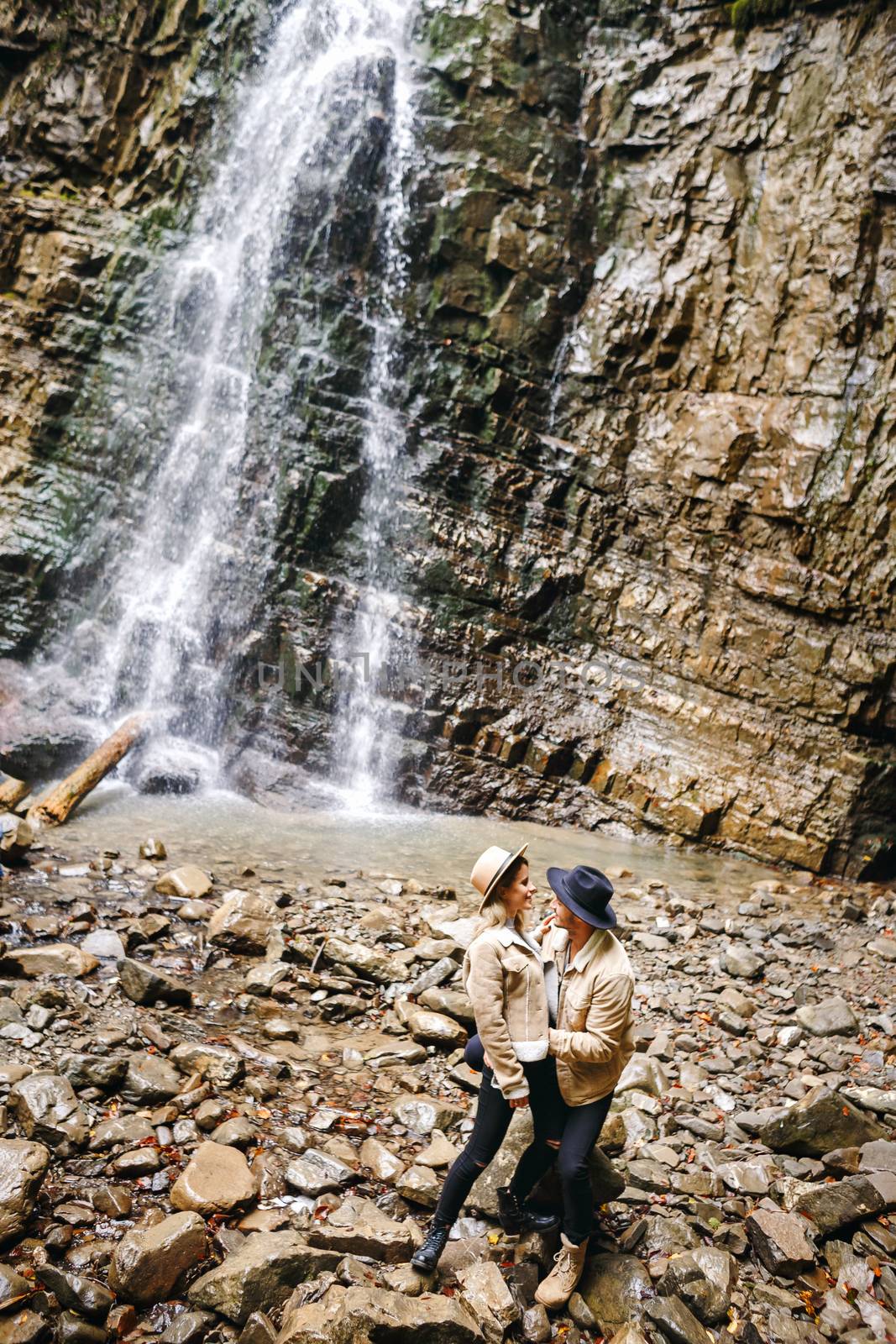 Young and beautiful couple at the mountain waterfall - Happy tourists visiting mountains. Lovestory. Tourists in hats. Military fashion