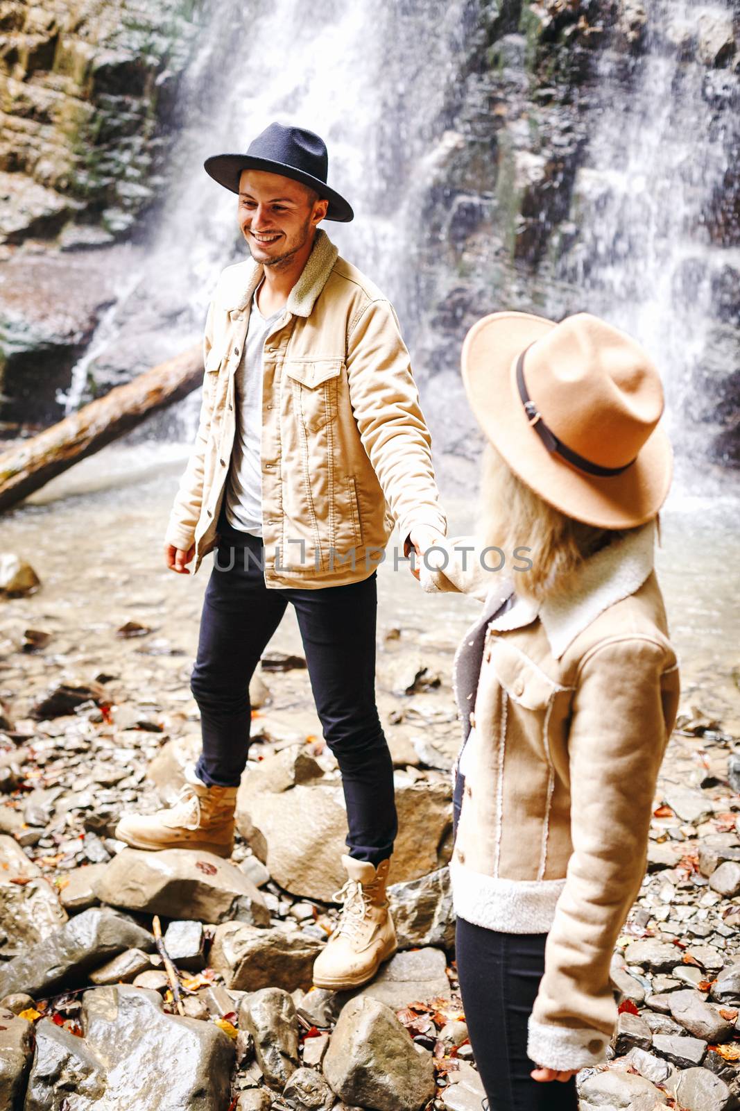 Young and beautiful couple at the mountain waterfall - Happy tourists visiting mountains. Lovestory. Tourists in hats. Military fashion