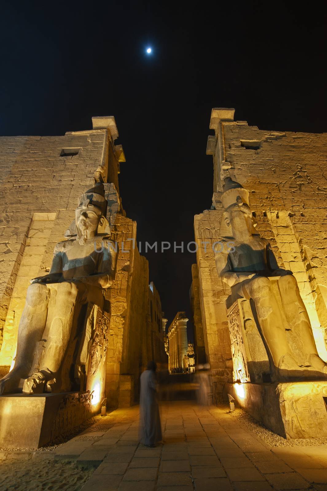 Large statues of Ramses II at entrance pylon to ancient egyptian Luxor Temple lit up during night